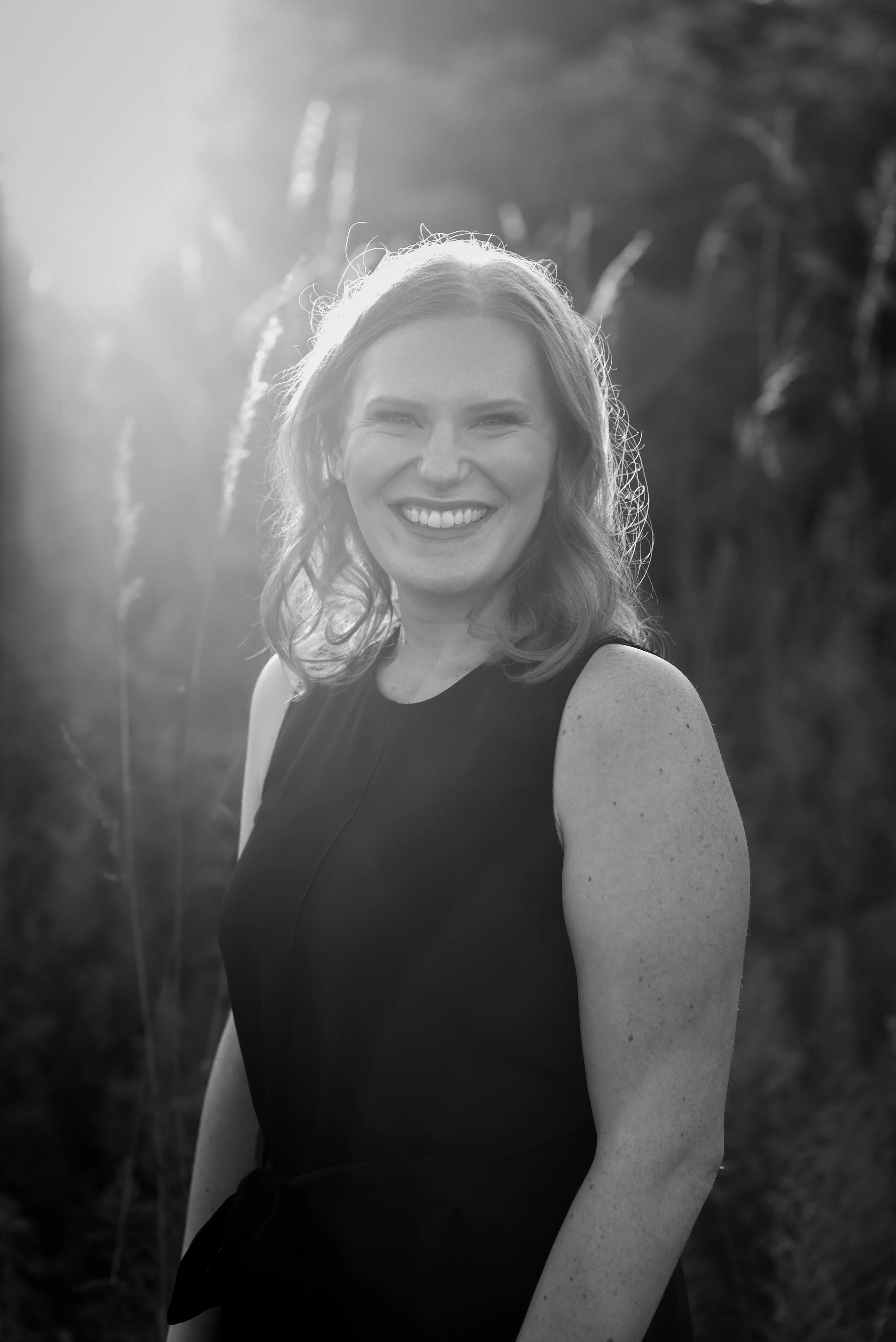 Black and white portrait of a woman smiling outdoors with sunlight behind her.