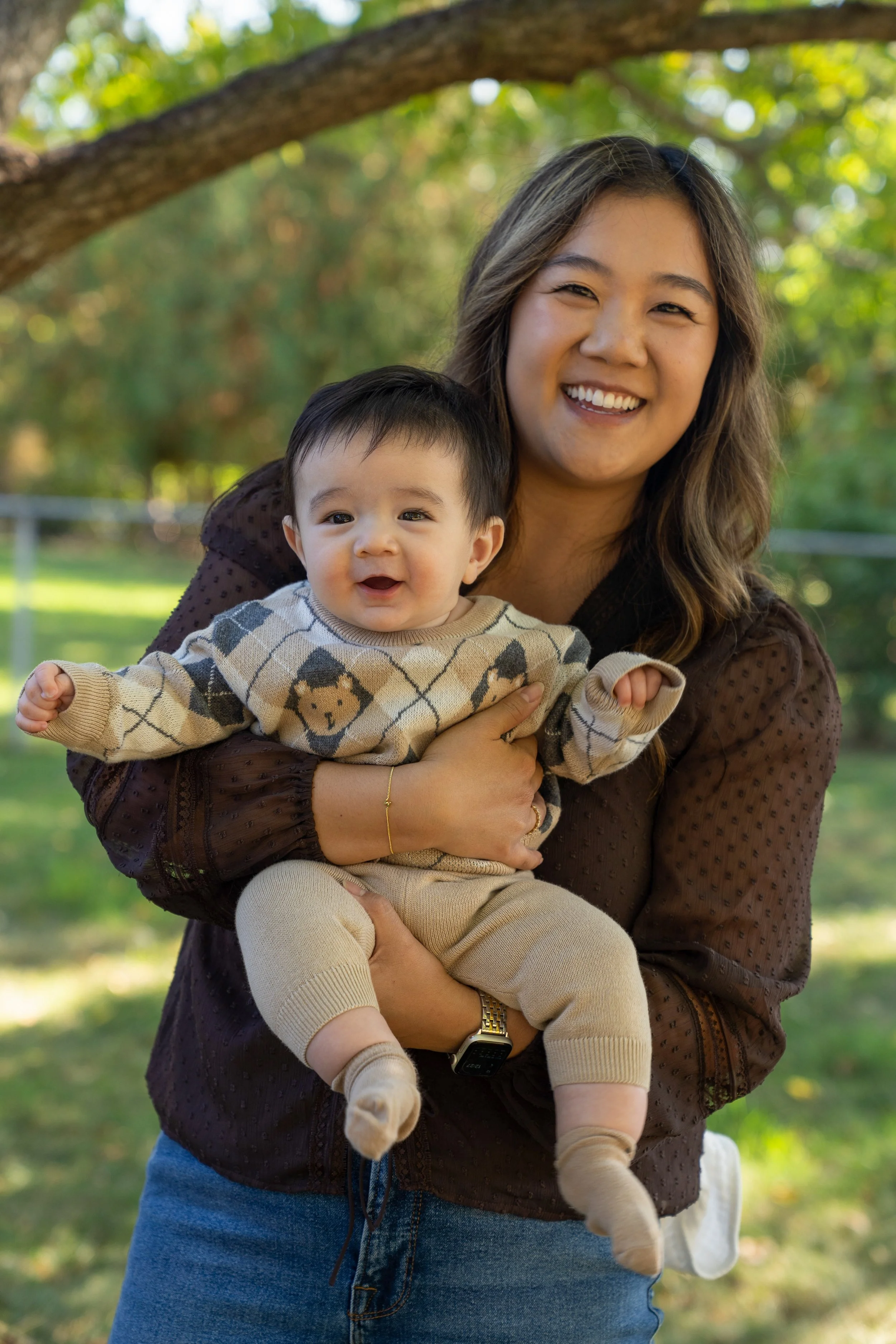 A smiling young woman holding a joyful baby outdoors in a park with trees and greenery in the background.