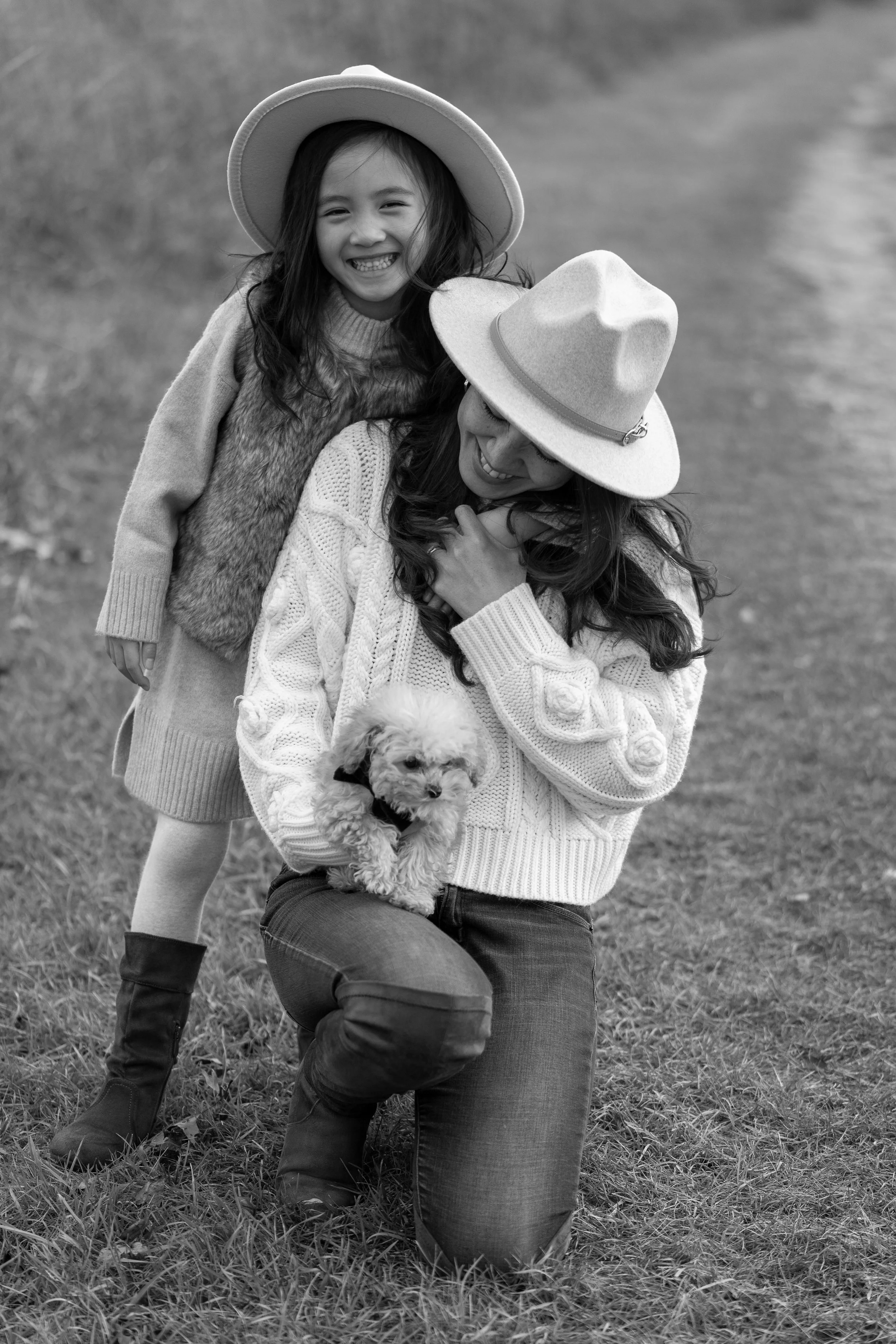 A woman kneeling on the grass with a small puppy in her arms, a young girl standing behind her, both wearing hats and smiling, in an outdoor setting.