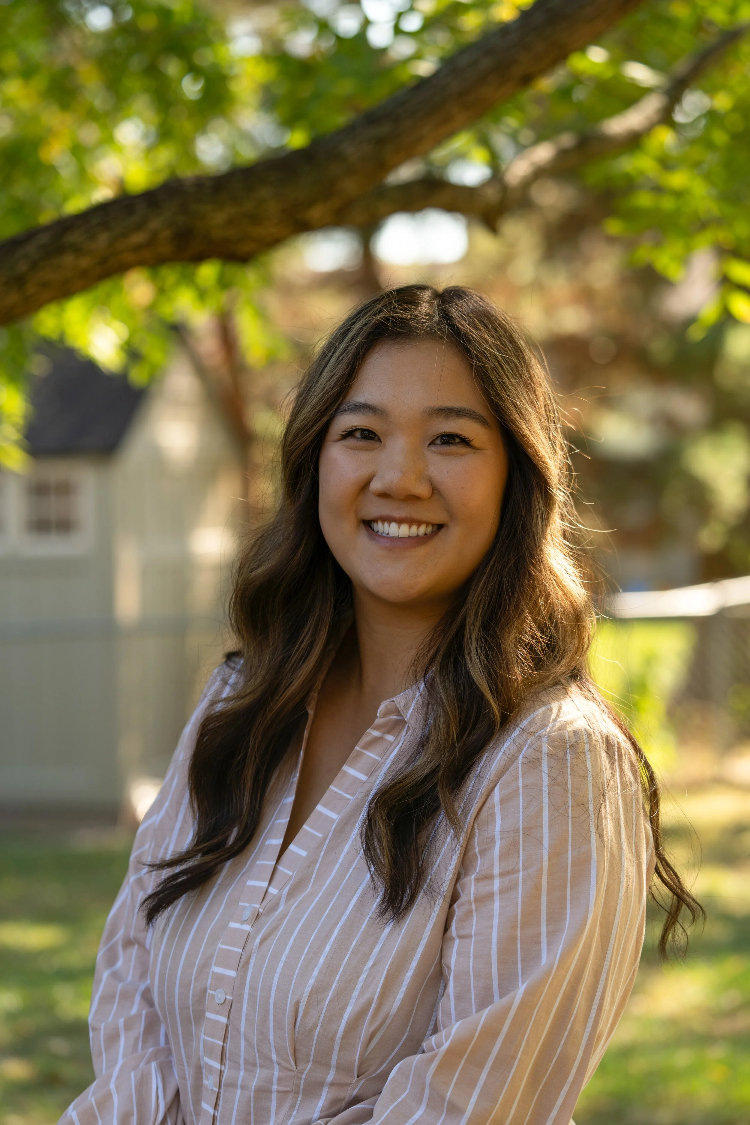 A woman with long wavy hair smiling outdoors with trees and houses in the background.