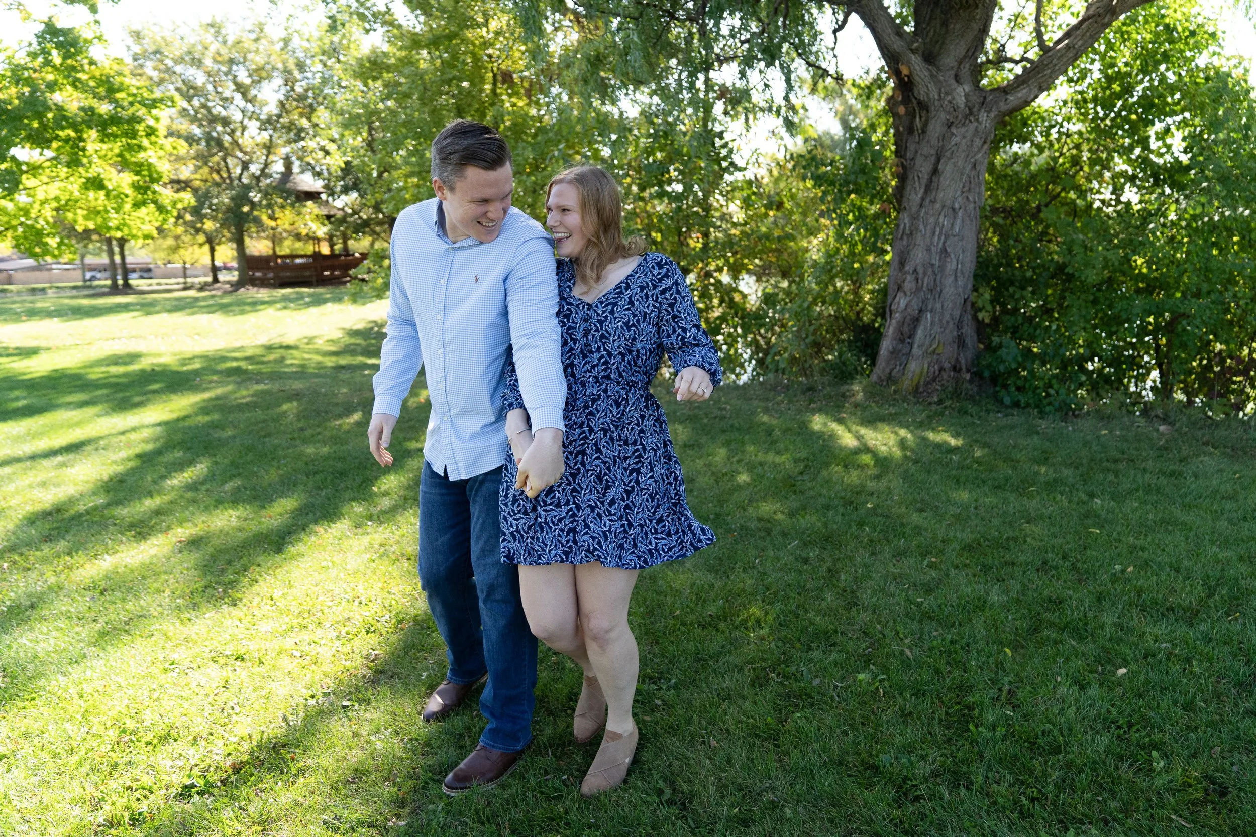 A smiling young couple playing and holding hands in a grassy park with trees.