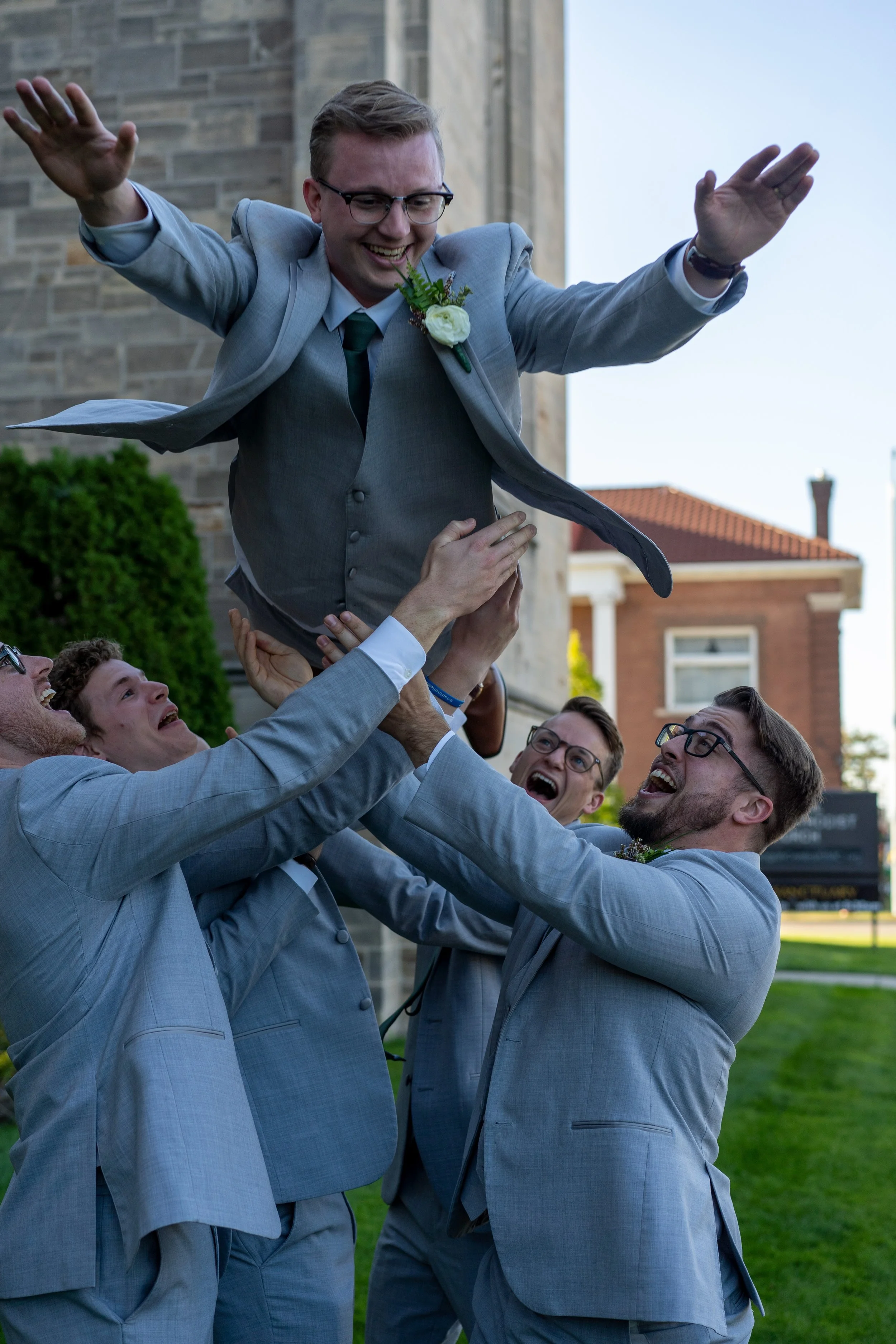 A group of five groomsmen lifting a smiling groom in a gray suit and glasses, outdoors in front of a stone building. They are celebrating at a wedding.