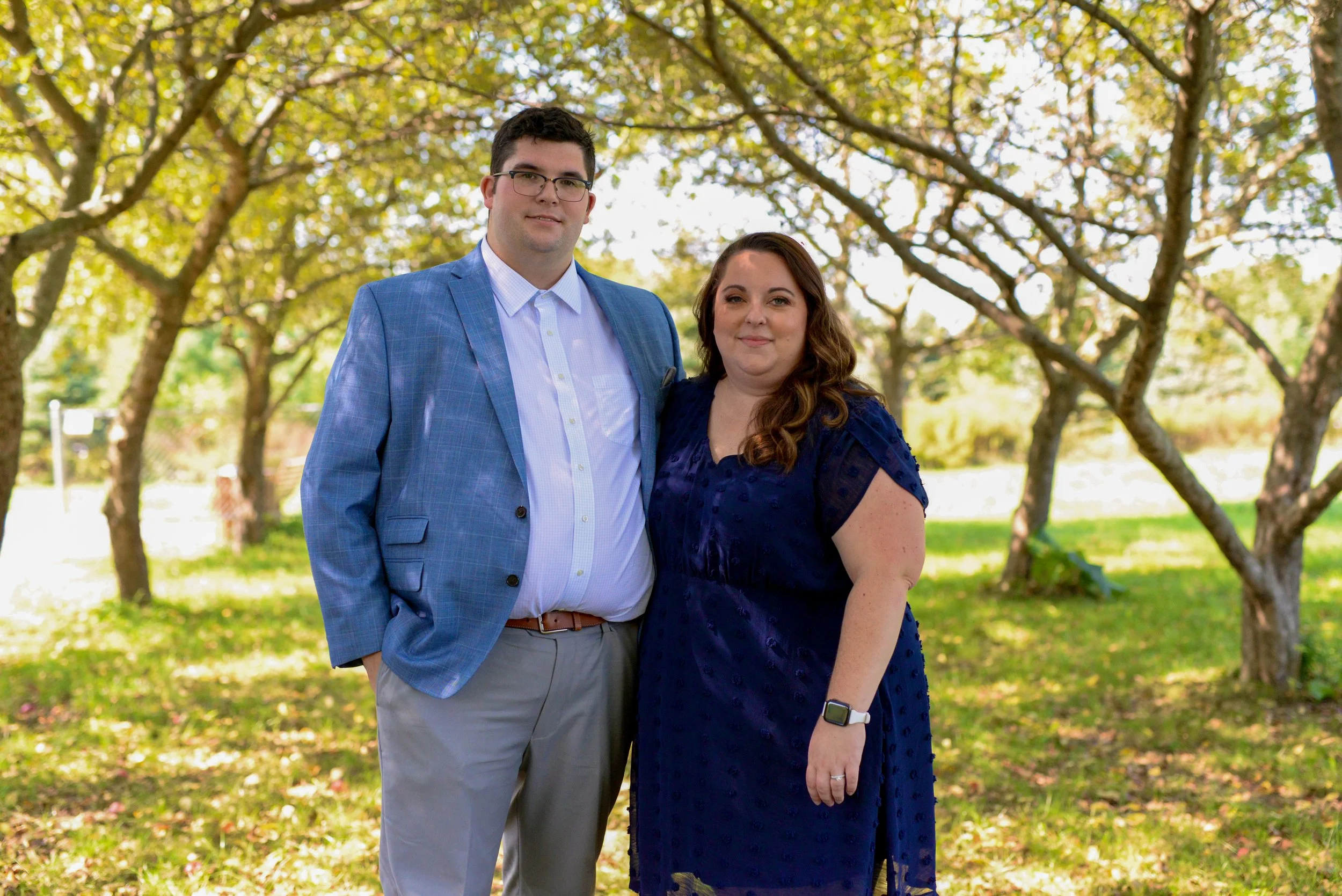 A man and woman standing outdoors in a park with trees, dressed in formal attire. The man wears a blue suit jacket, white shirt, and beige pants. The woman wears a dark blue dress and a smartwatch. They are smiling and close together.