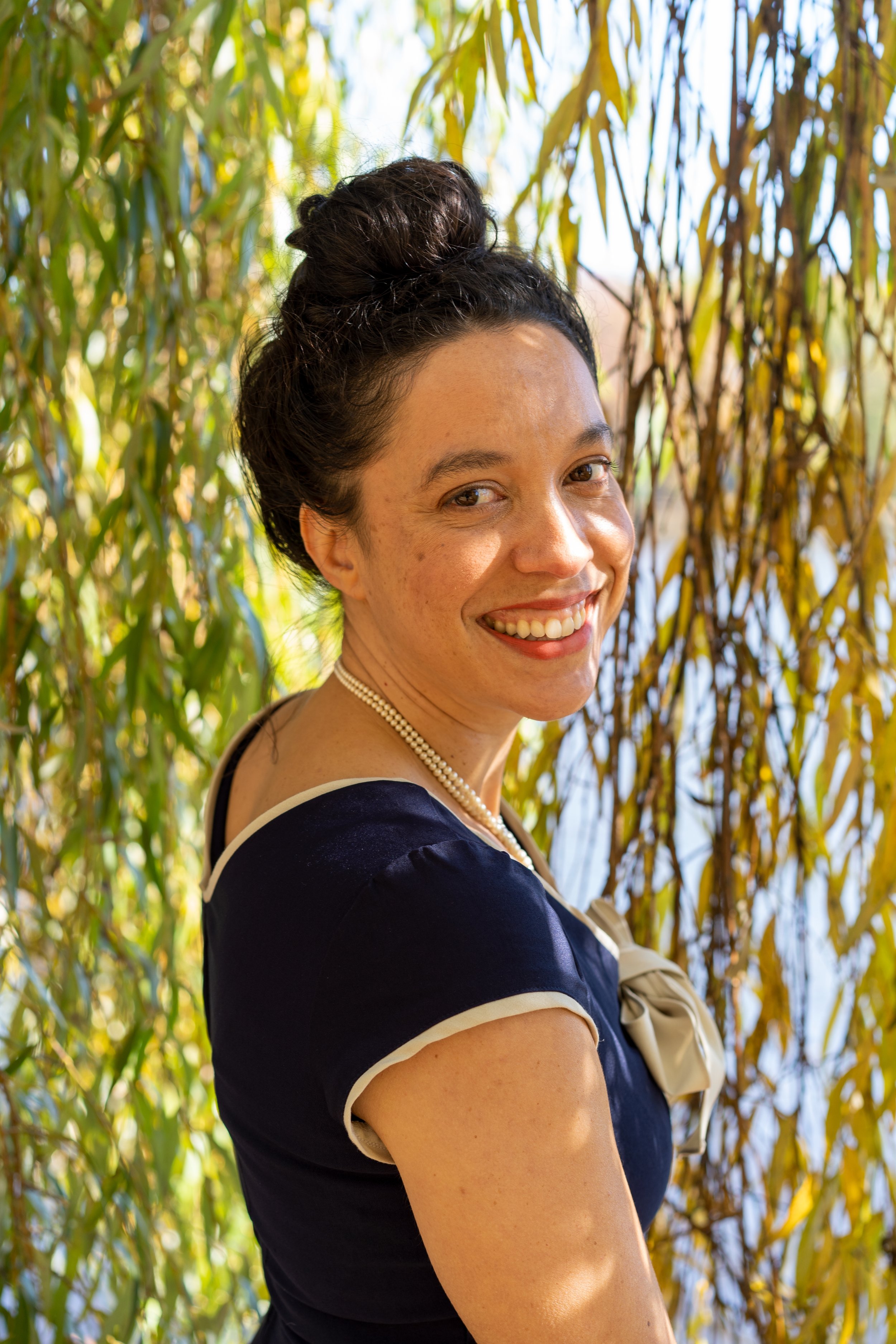 A woman with dark hair styled in a bun, wearing a black dress with beige trim and a pearl necklace, stands outdoors among yellow and green foliage, smiling at the camera.