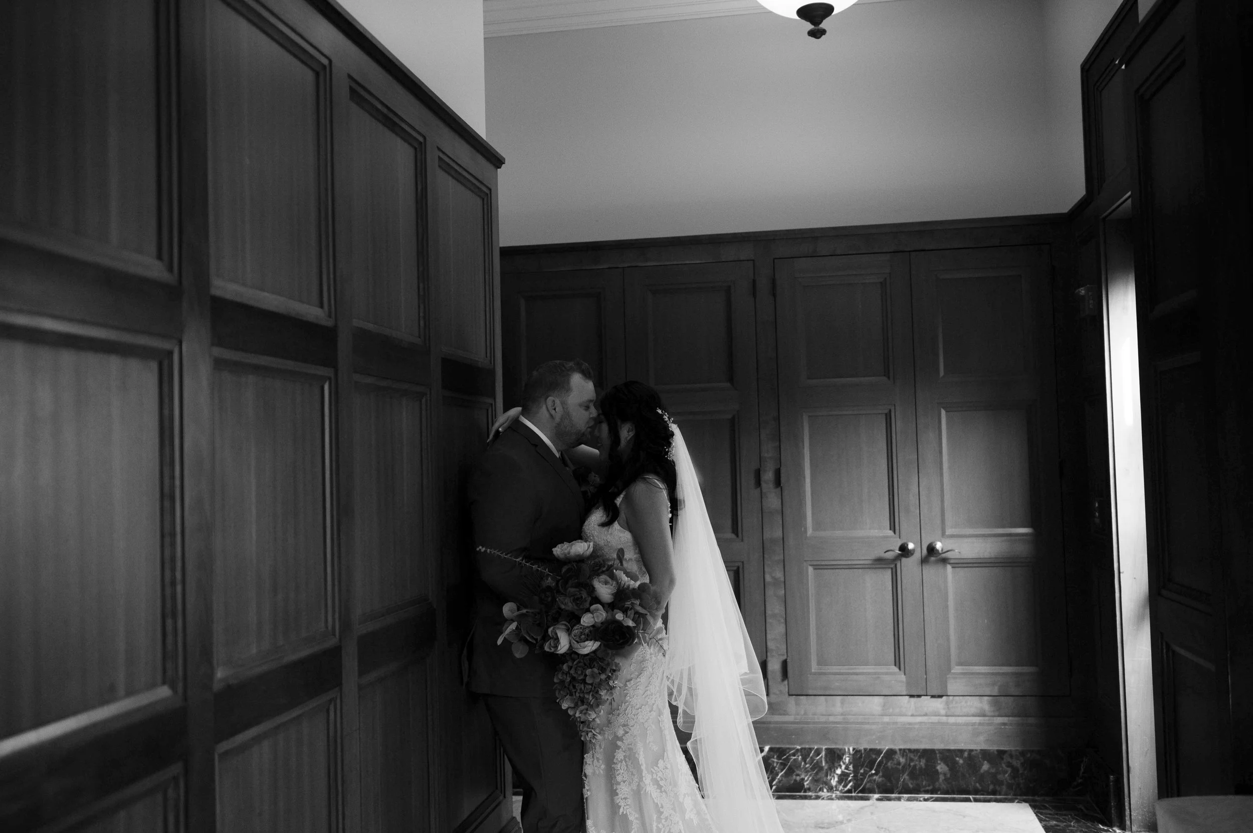 A black and white photograph of a bride and groom sharing an intimate moment in a room with wood-paneled walls. The bride is wearing a lace wedding dress and veil, holding a large bouquet, while the groom is dressed in a suit. They are standing close