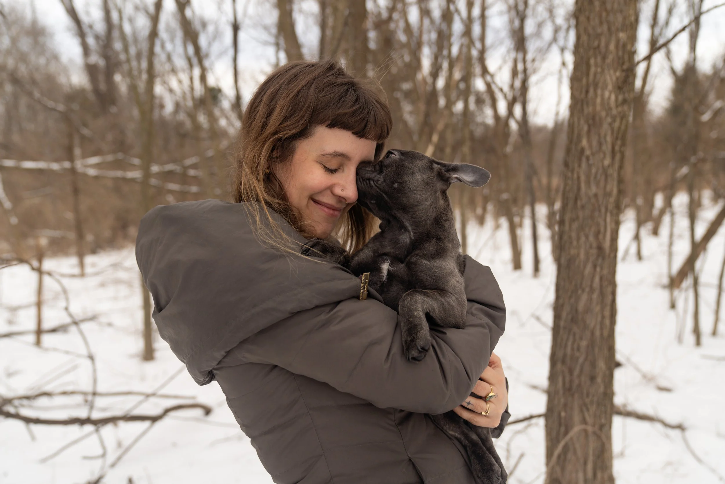 A woman holding a dark puppy in a snowy wooded area, smiling with her eyes closed as the puppy sniffs her face.