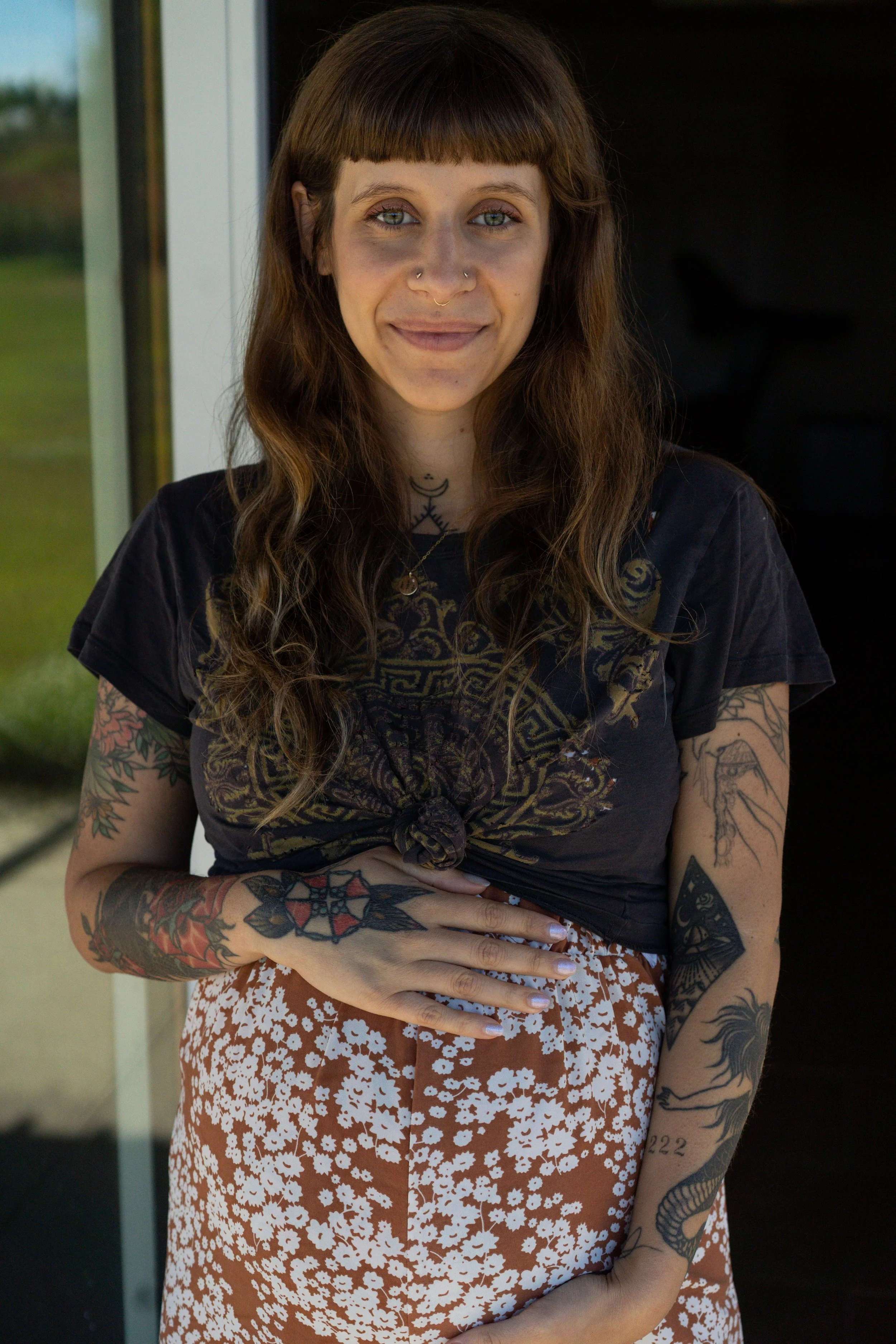 A woman with long wavy brown hair, tattoos on her arms, and multiple facial piercings, standing indoors in front of a glass door with a green outdoor landscape visible behind her.