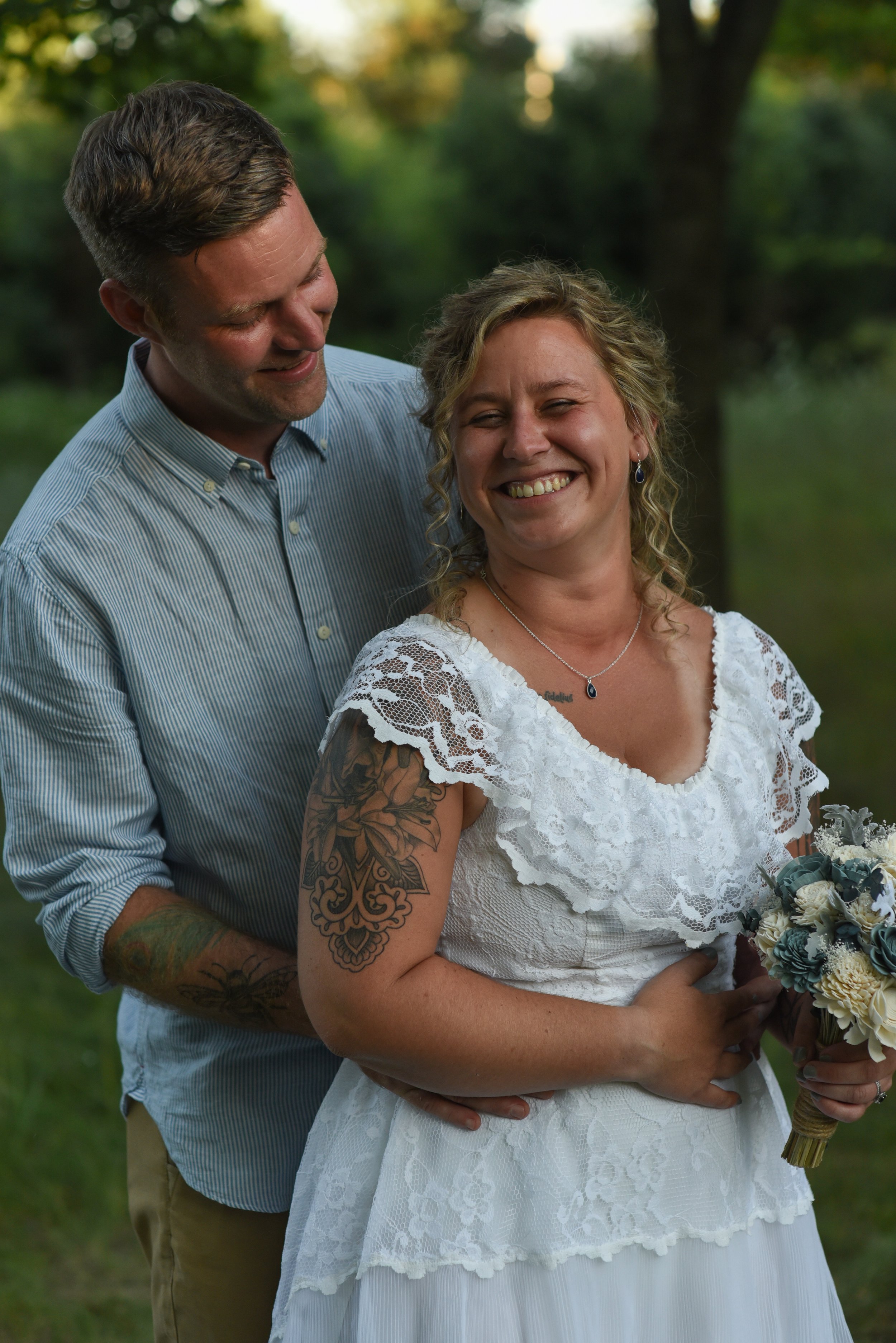 A smiling woman in a white lace dress holding a bouquet, with a man in a striped shirt standing behind her, outdoors in a green, wooded area.