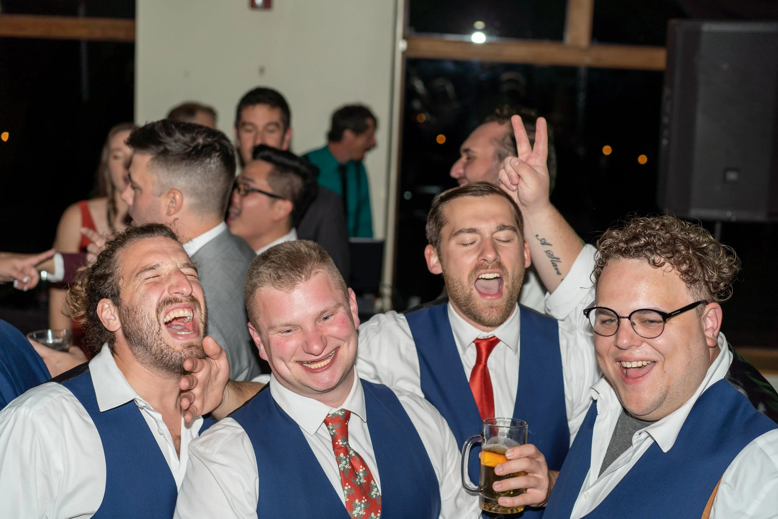 Group of four men wearing blue vests and white shirts, laughing and having fun at a party, one holding a drink, with people dancing and celebrating in the background.