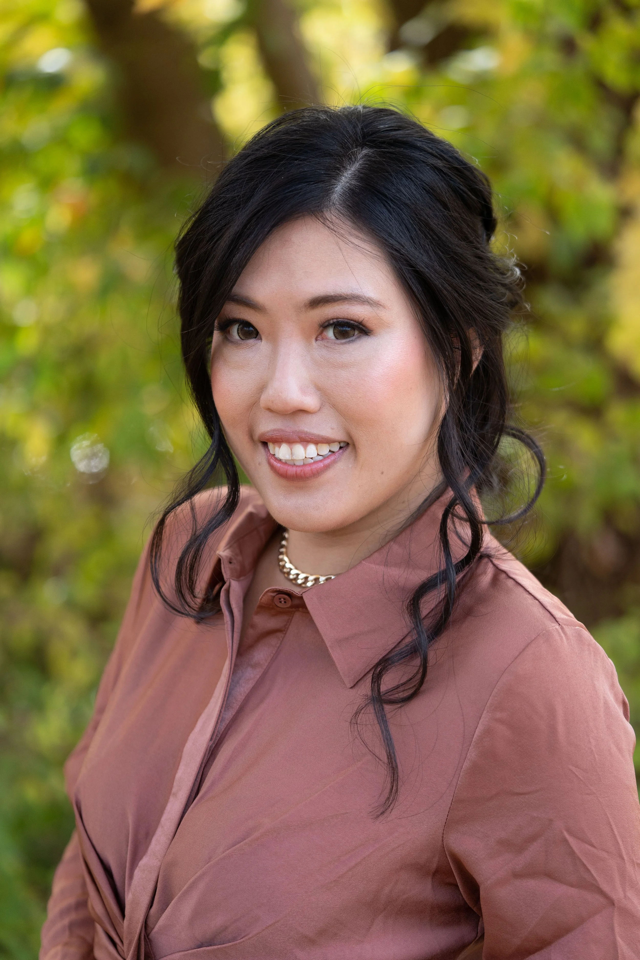 A woman with dark hair styled in loose waves, wearing a pinkish-brown blouse and a pearl necklace, smiling outdoors with green foliage in the background.
