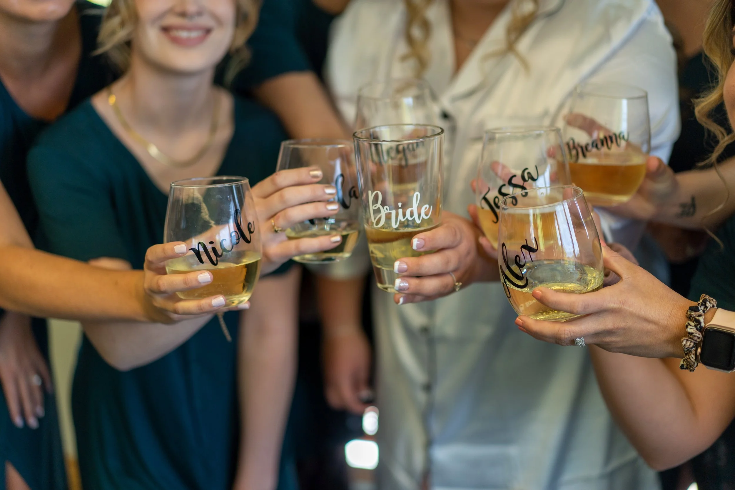 Group of women holding glasses of champagne with personalized labels for a wedding celebration, some glasses say 'Nicole', 'Bride', 'Jesse', and 'Alex'.
