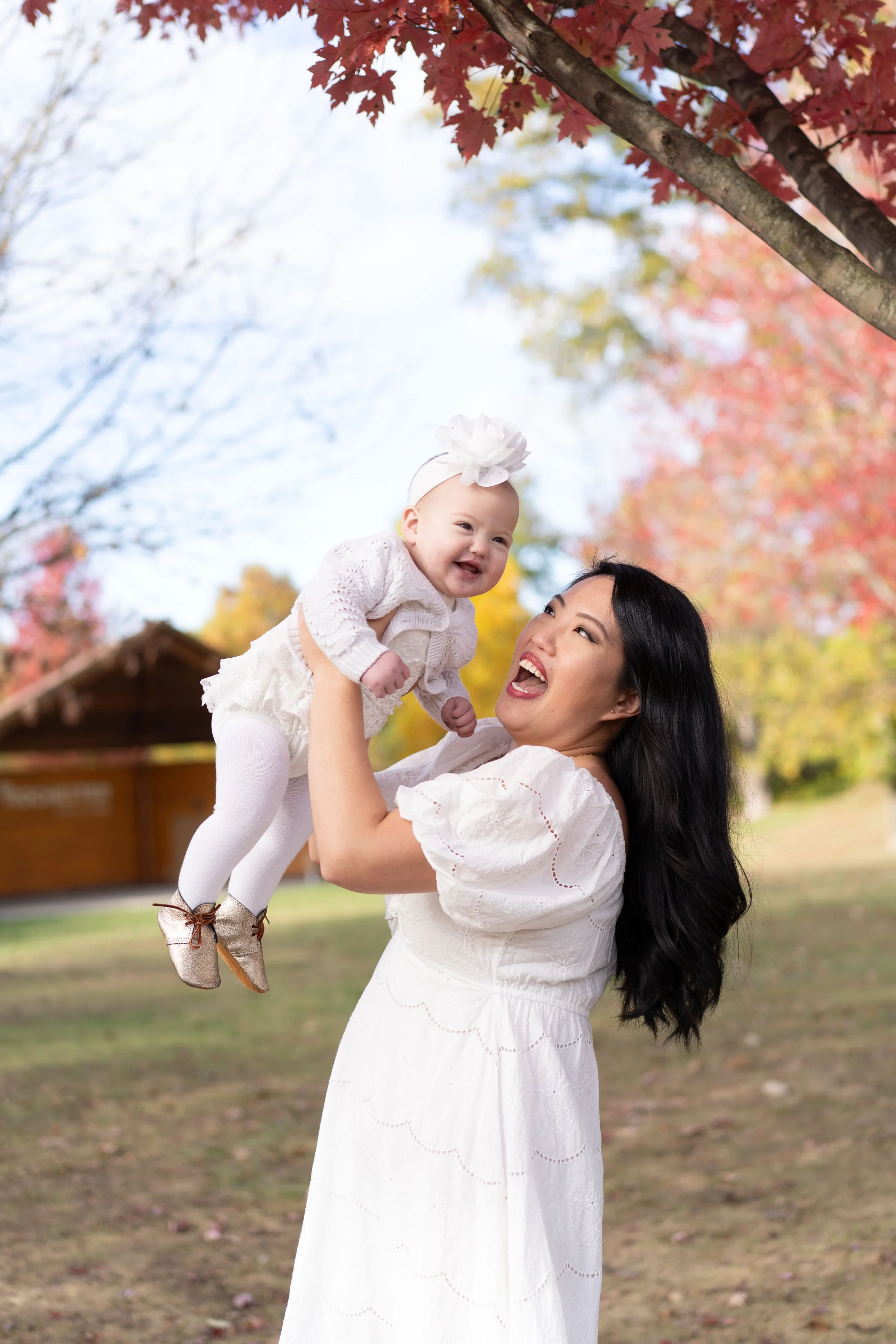 A woman lifting a smiling baby girl outside in a park with autumn-colored trees.