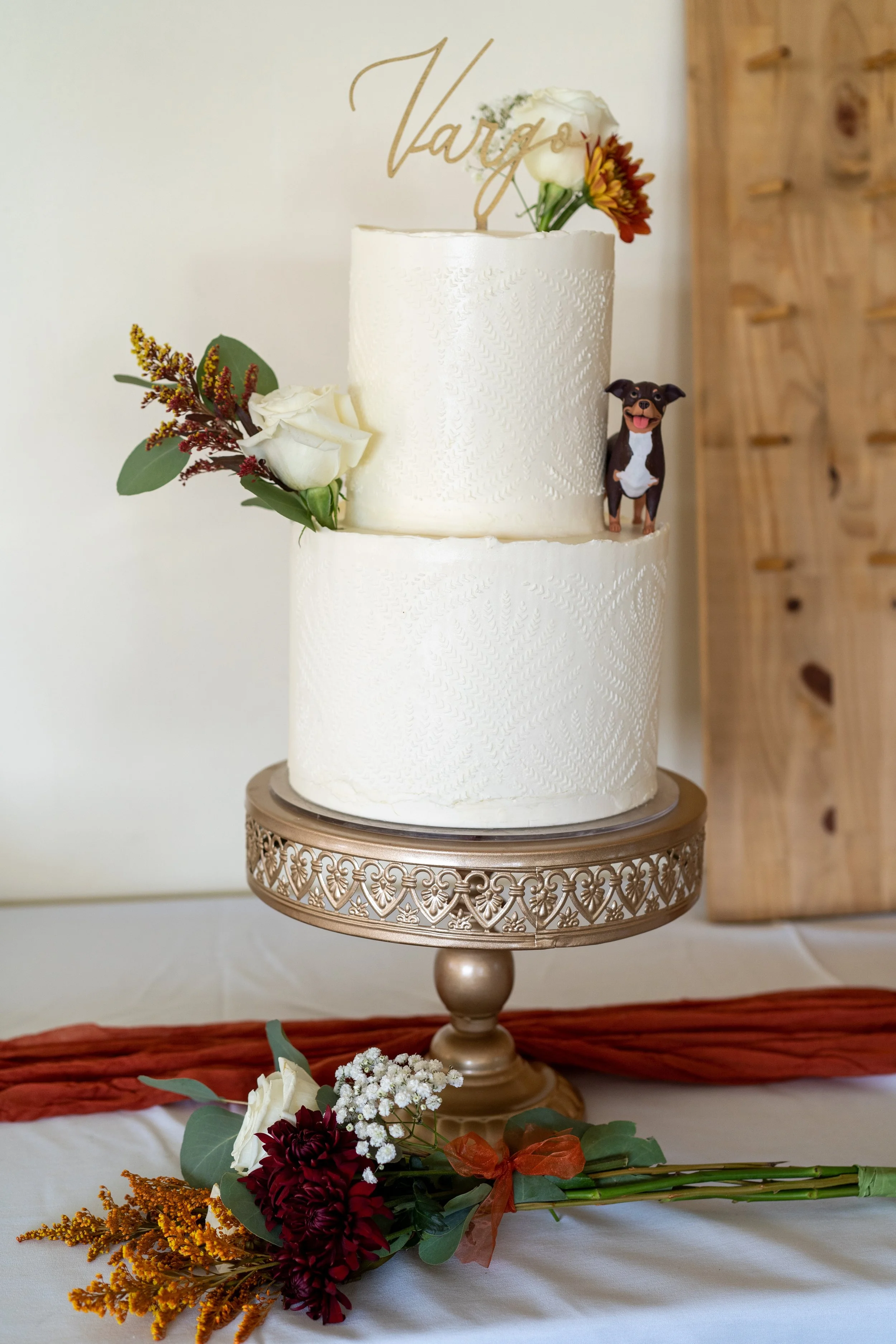A two-tier wedding cake decorated with white flowers, greenery, and a cake topper that says 'Vargo.' The cake is on a decorative gold cake stand, with a bouquet of flowers including white roses, red and burgundy blooms, and greenery in front of it, a
