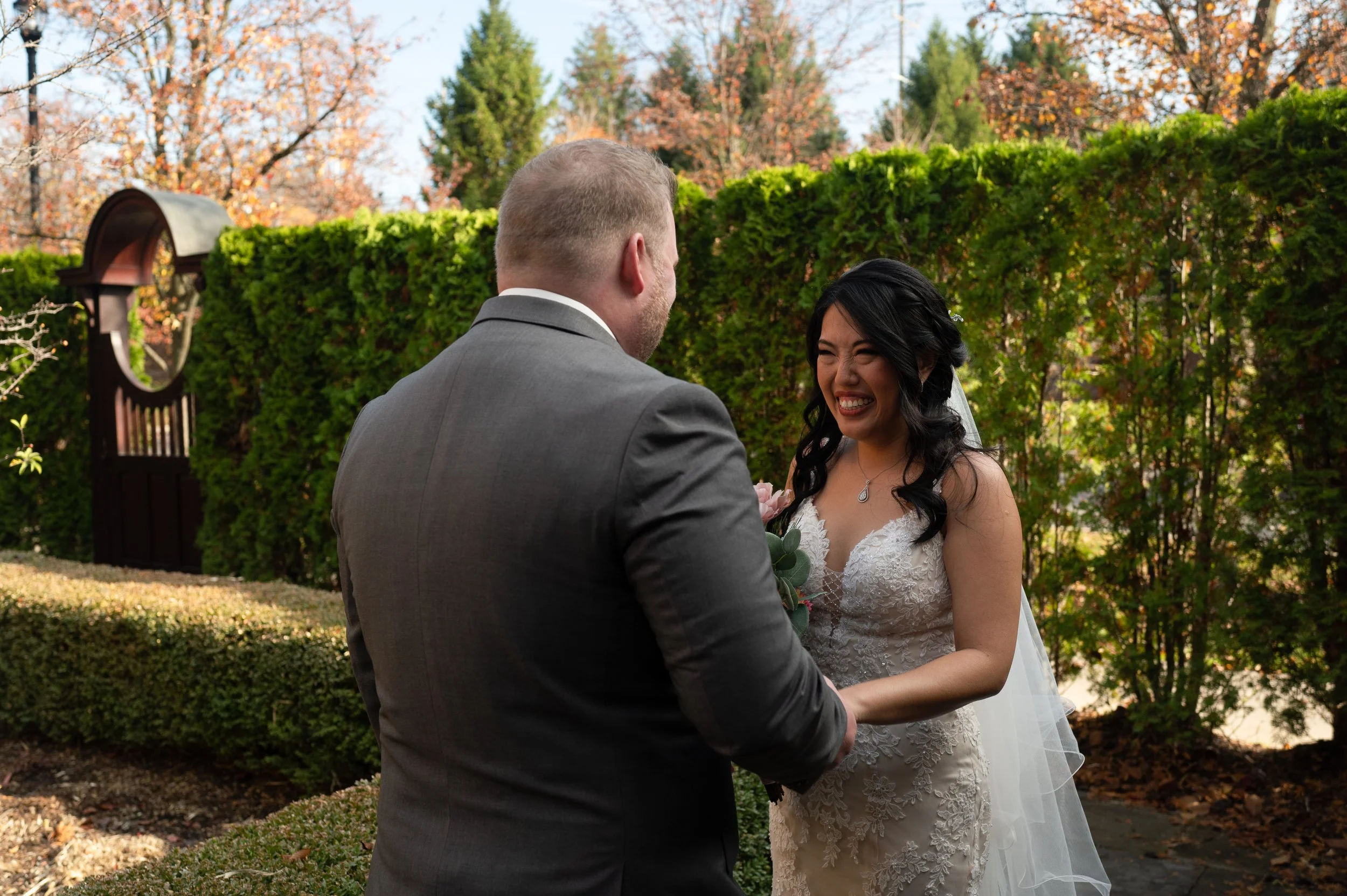 A bride and groom are holding hands and smiling at each other outdoors during a wedding ceremony on a sunny day with autumn foliage in the background.