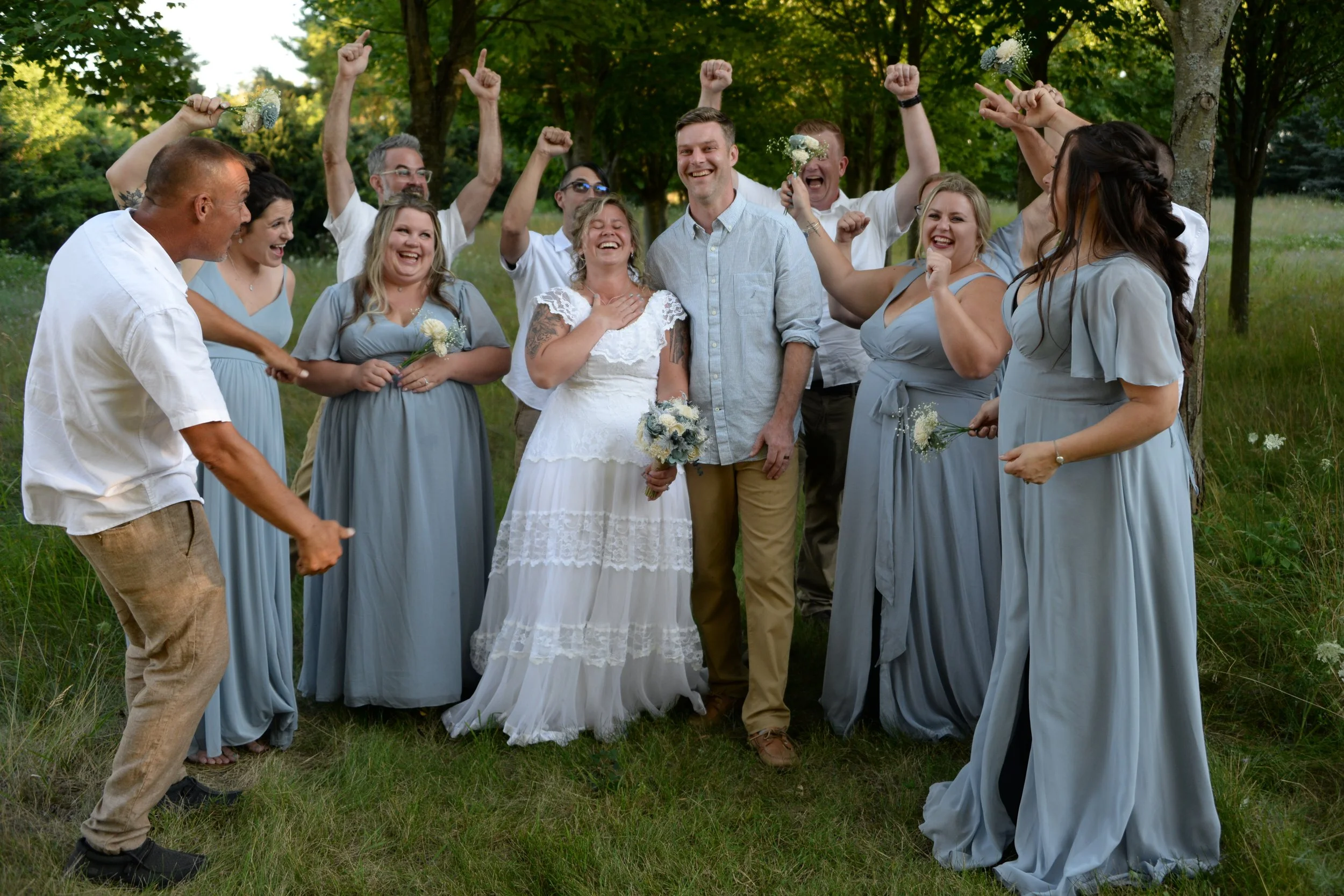 A wedding celebration outdoors with the bride and groom surrounded by friends and family. Everyone is smiling and raising their fists or holding flowers.
