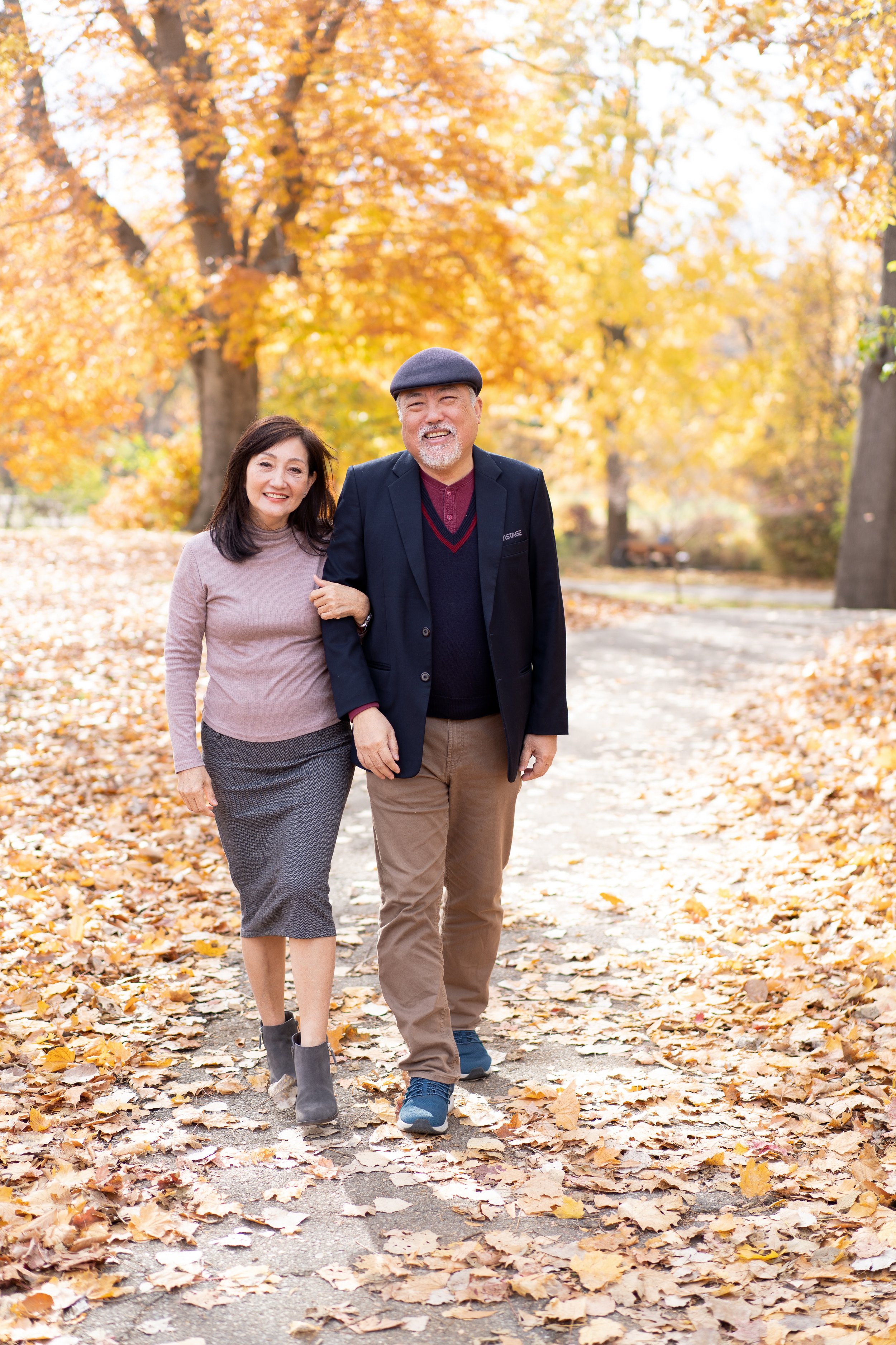 A smiling middle-aged couple walking arm in arm on a leaf-covered park path during autumn, with orange and yellow trees in the background.