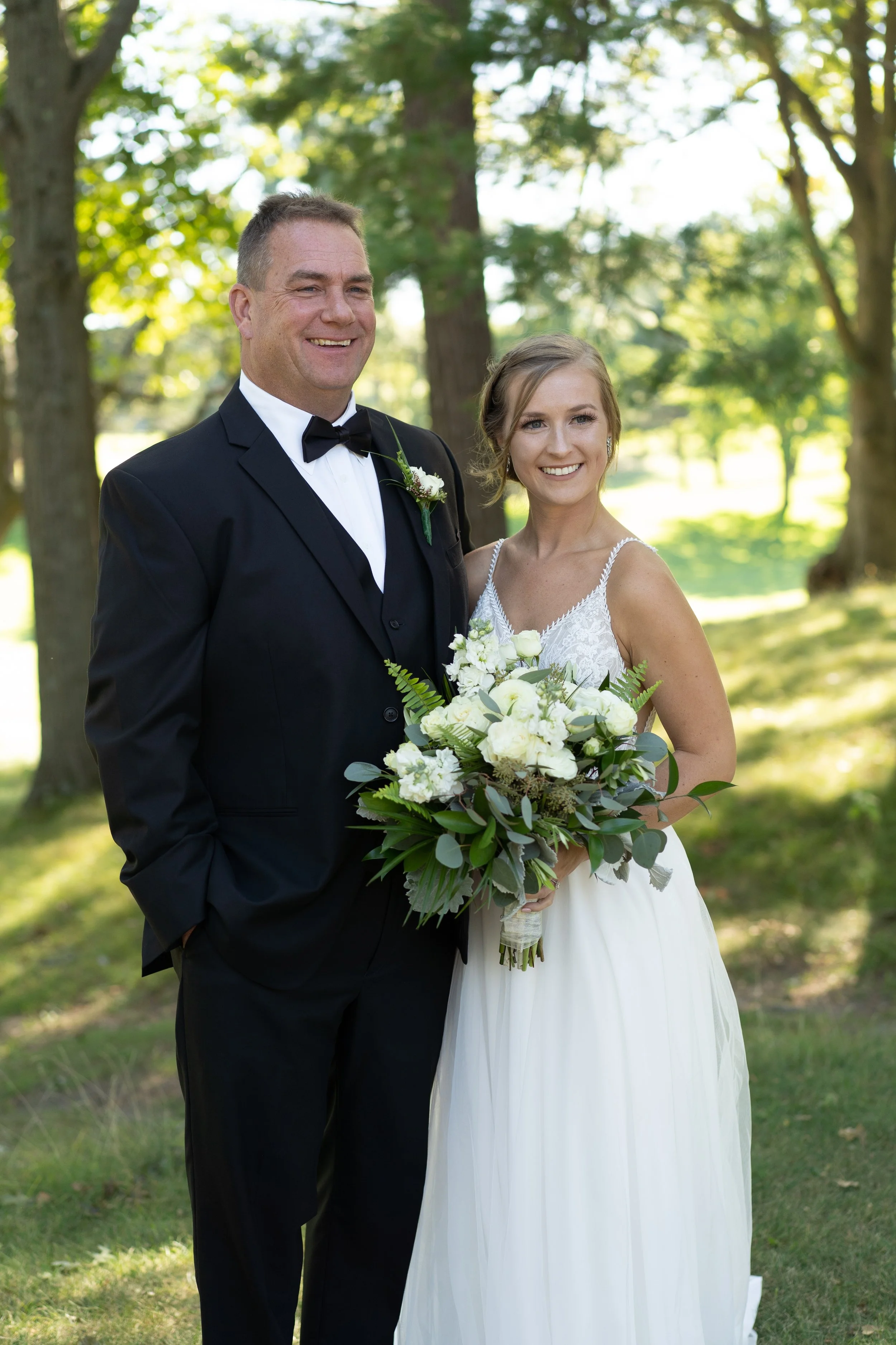 A bride and a groom standing outdoors in a wooded area on their wedding day, both smiling. The bride is in a white wedding dress holding a bouquet of white flowers and greenery. The groom is in a black tuxedo with a bow tie, dressed in a tuxedo with 