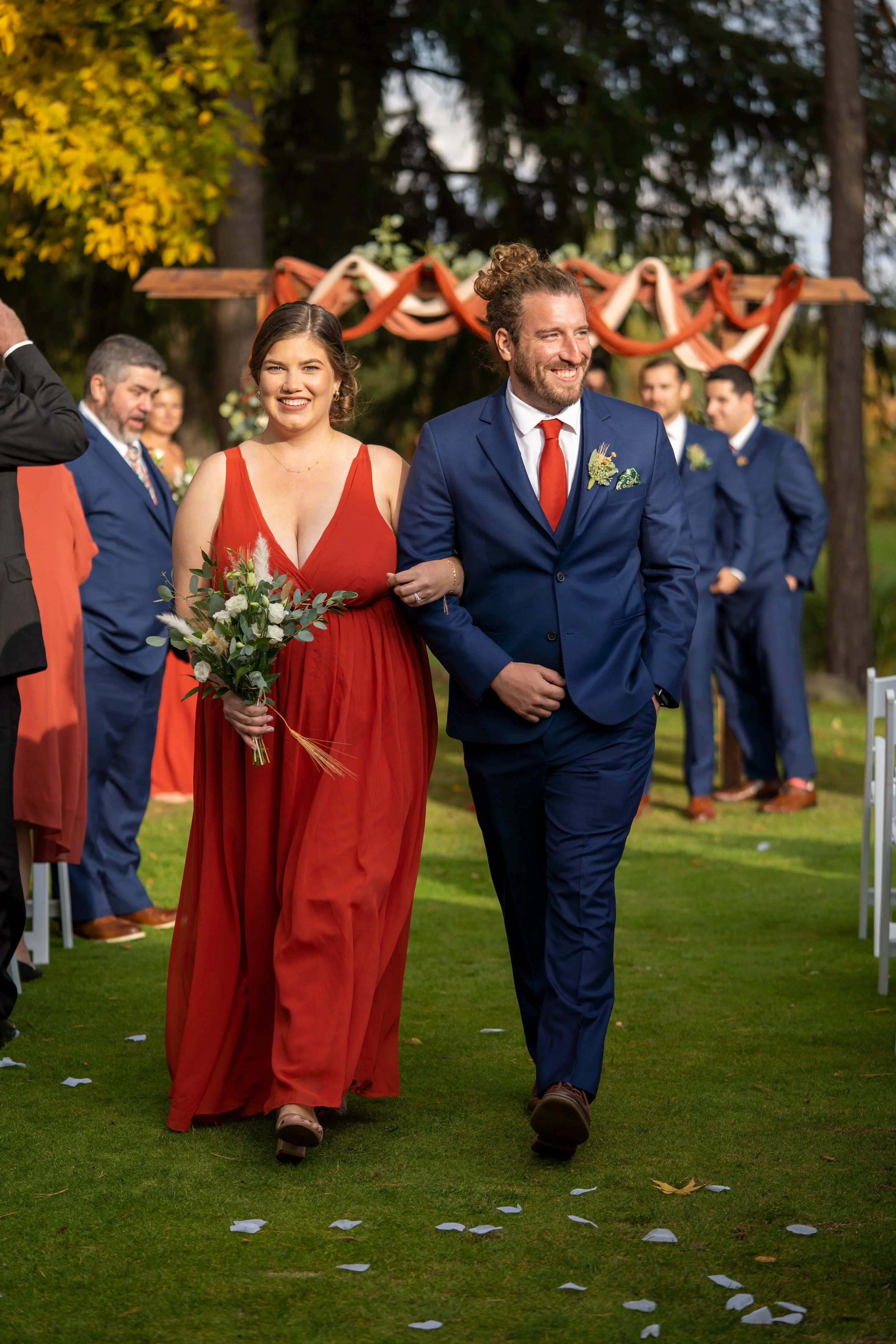 A couple walking arm-in-arm down an outdoor aisle at a wedding ceremony, with the bride holding a bouquet of flowers and smiling, and the groom smiling while in a blue suit. Guests are in the background, and a decorated arch is visible behind them.