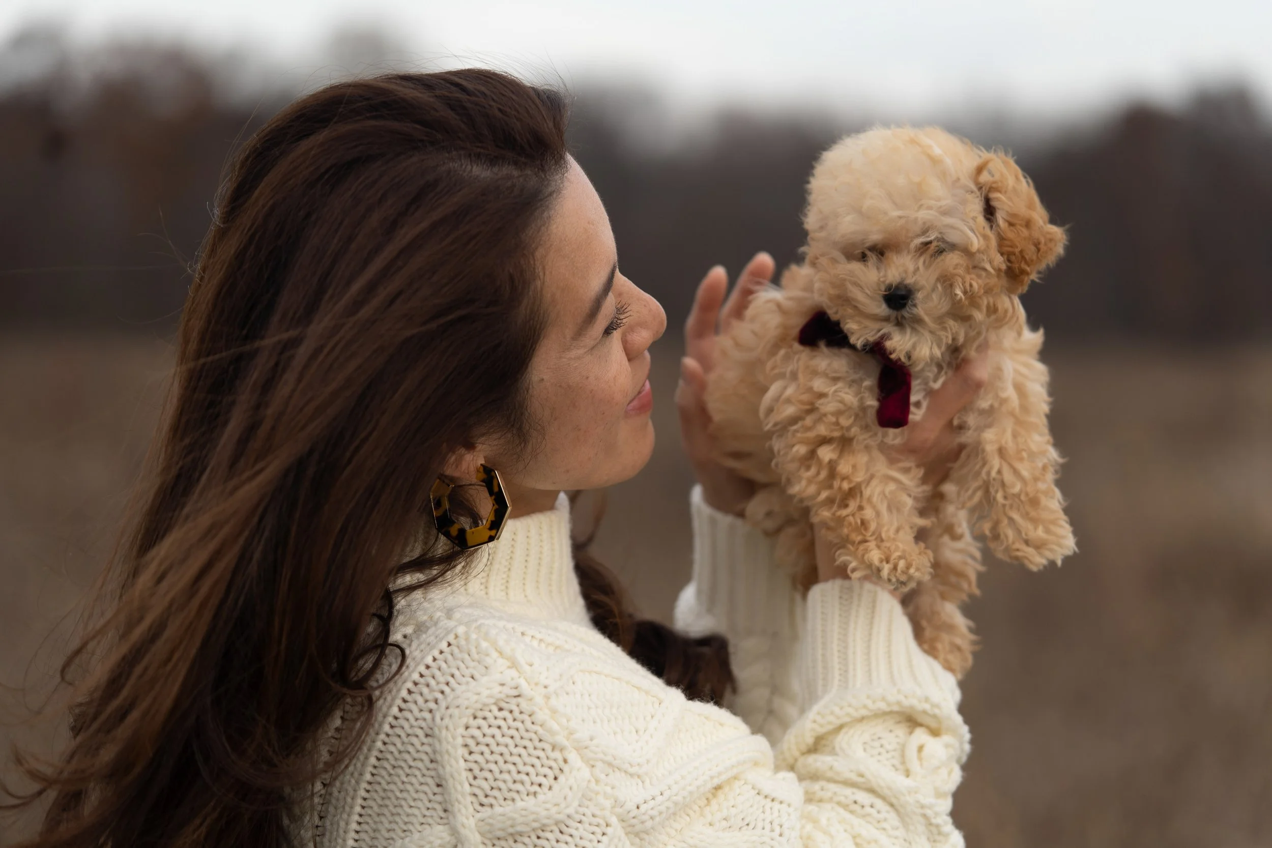 A woman with brown hair wearing a white sweater and large tortoiseshell earrings holds a small, fluffy, apricot-colored puppy with a maroon bow tie, outdoors with a blurred background.