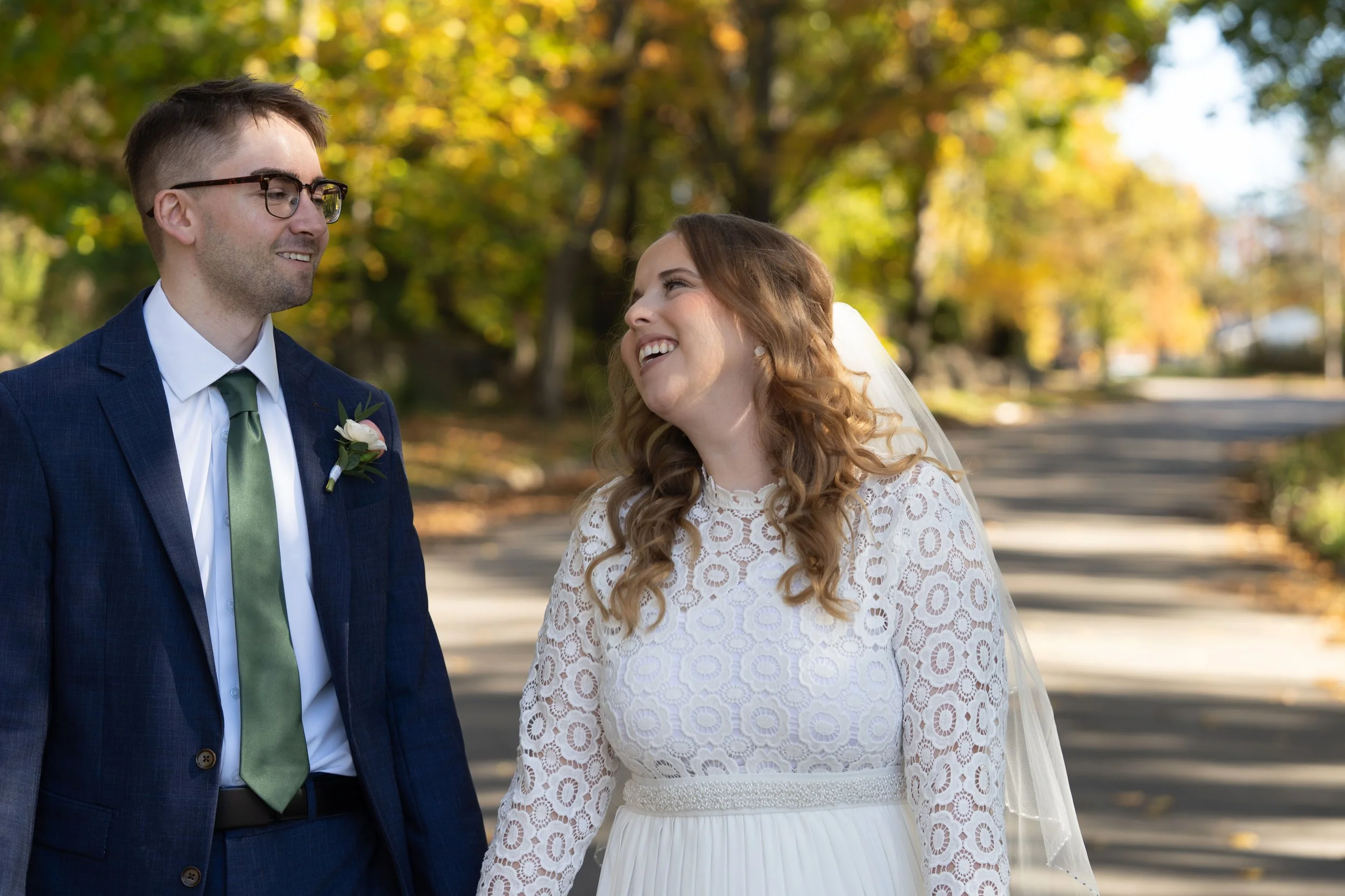 A newlywed couple walking outdoors in a park during autumn; the groom in a navy suit, white shirt, green tie, and glasses, and the bride in a white lace dress with long, curly hair and a veil, both smiling at each other.