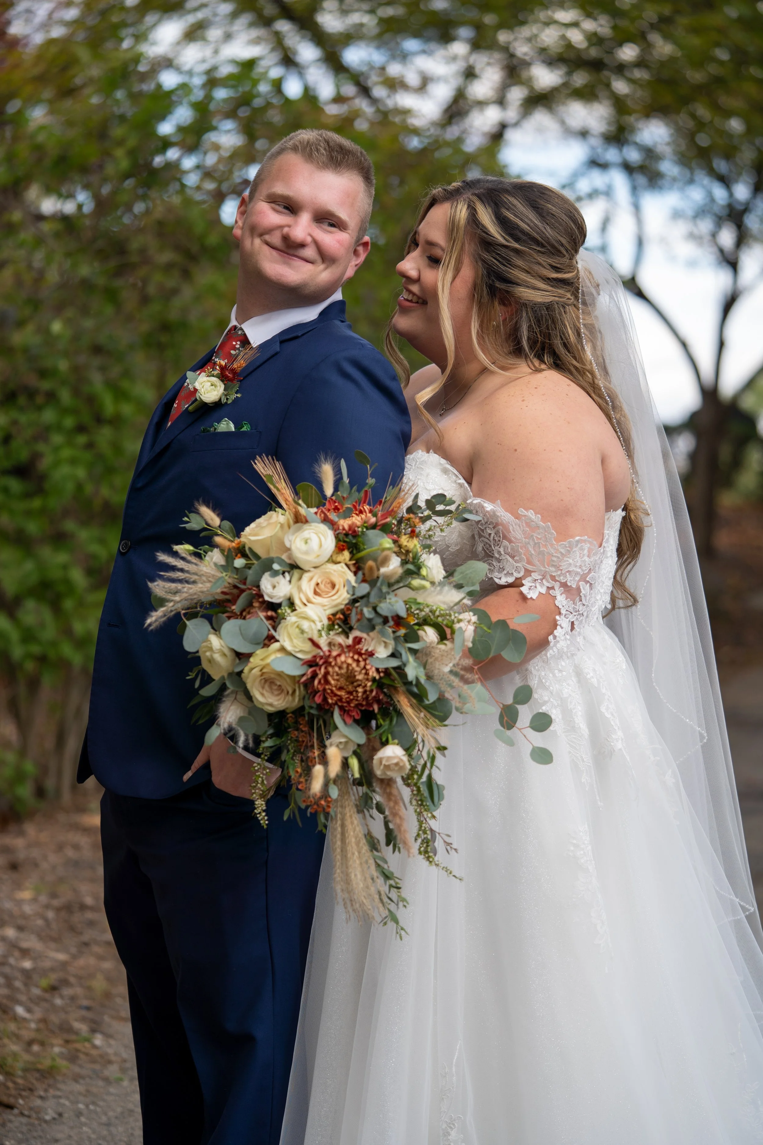 A bride and groom posing outdoors on their wedding day, with the bride holding a large bouquet of flowers and the groom dressed in a navy suit, trees in the background.