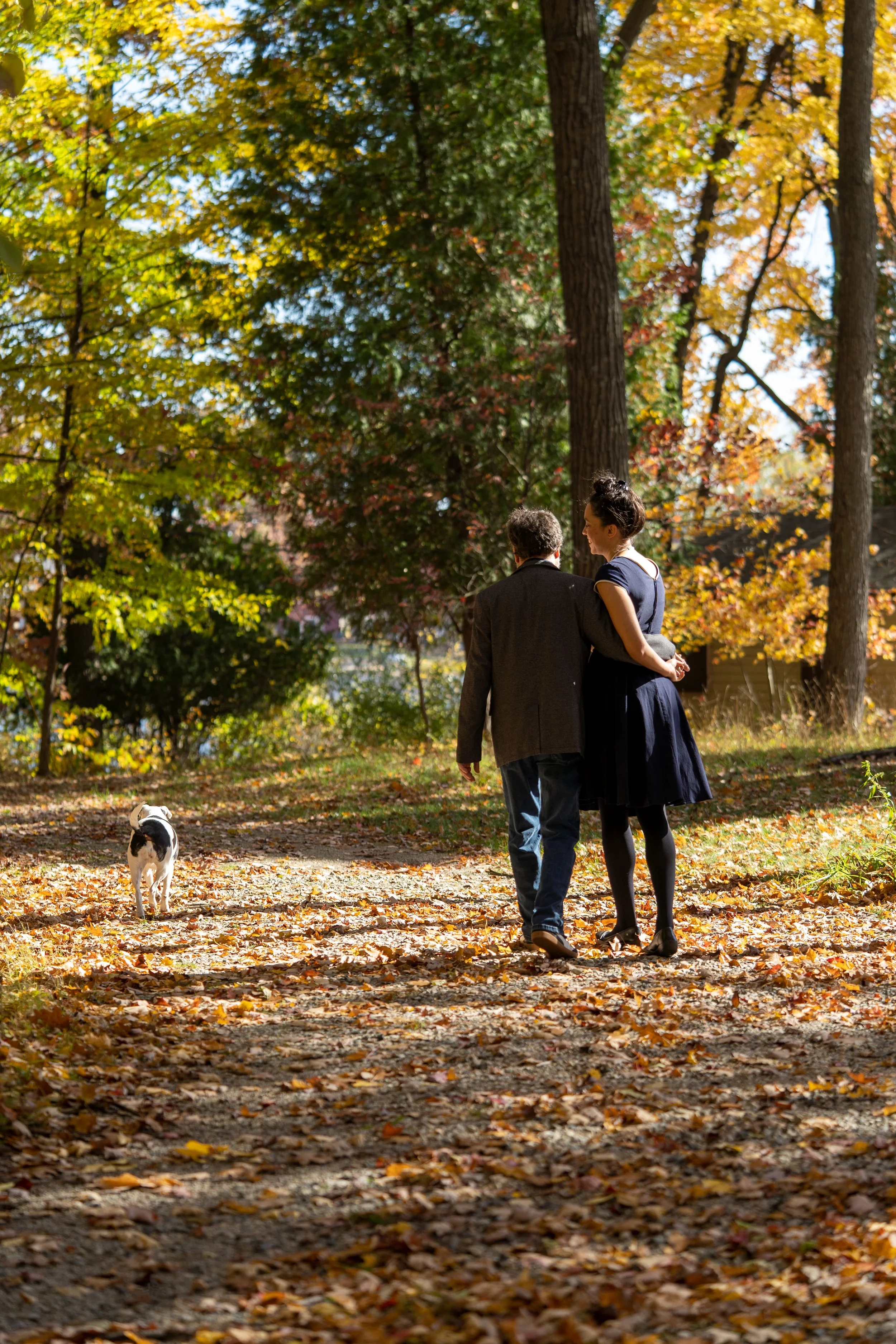 A couple walking together in a park during autumn, with a small white and black dog nearby, surrounded by colorful fall leaves and tall trees.