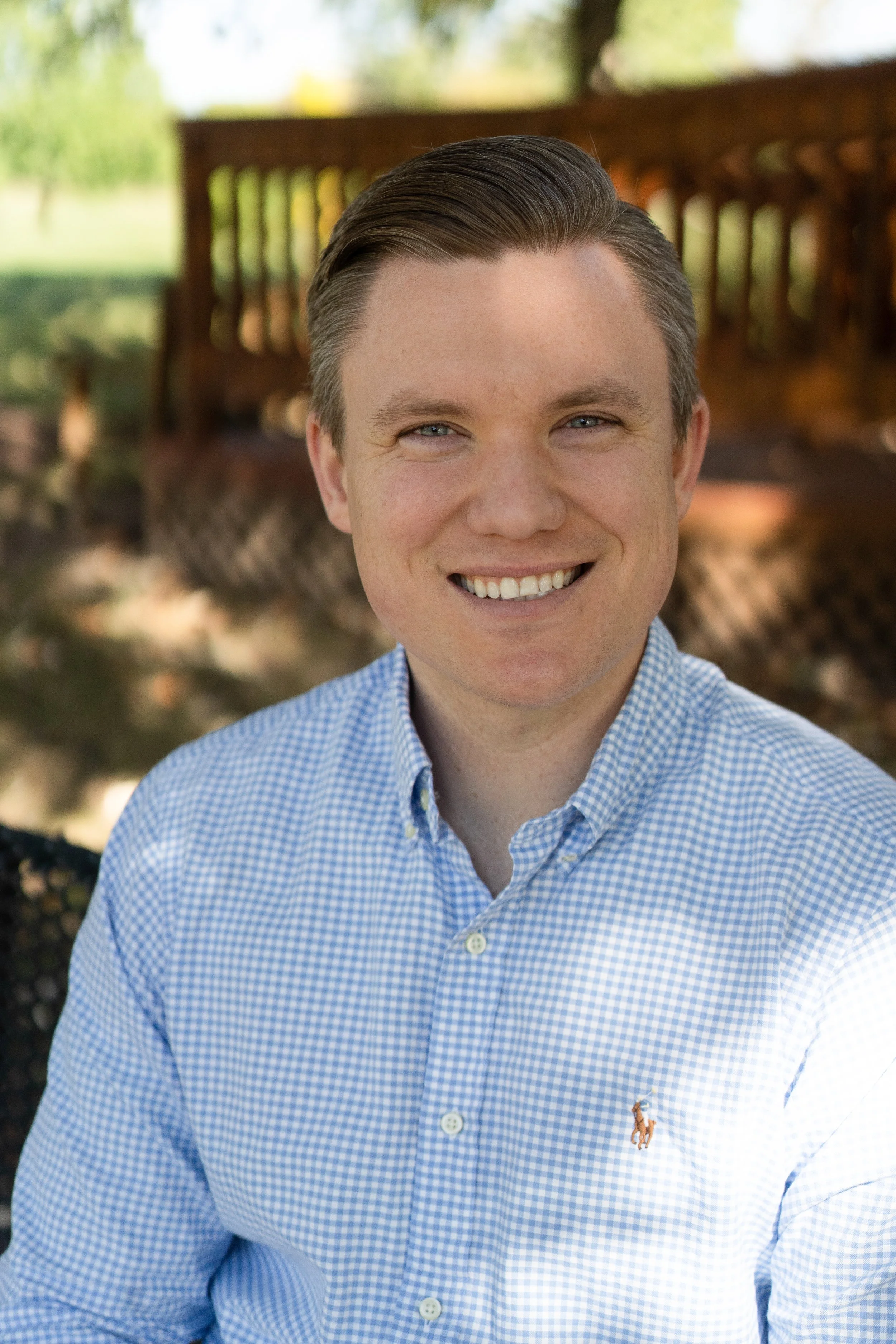 A young man with light skin and brown hair smiling outdoors, wearing a light blue checked button-up shirt, with a park or garden background featuring trees and a wooden fence.