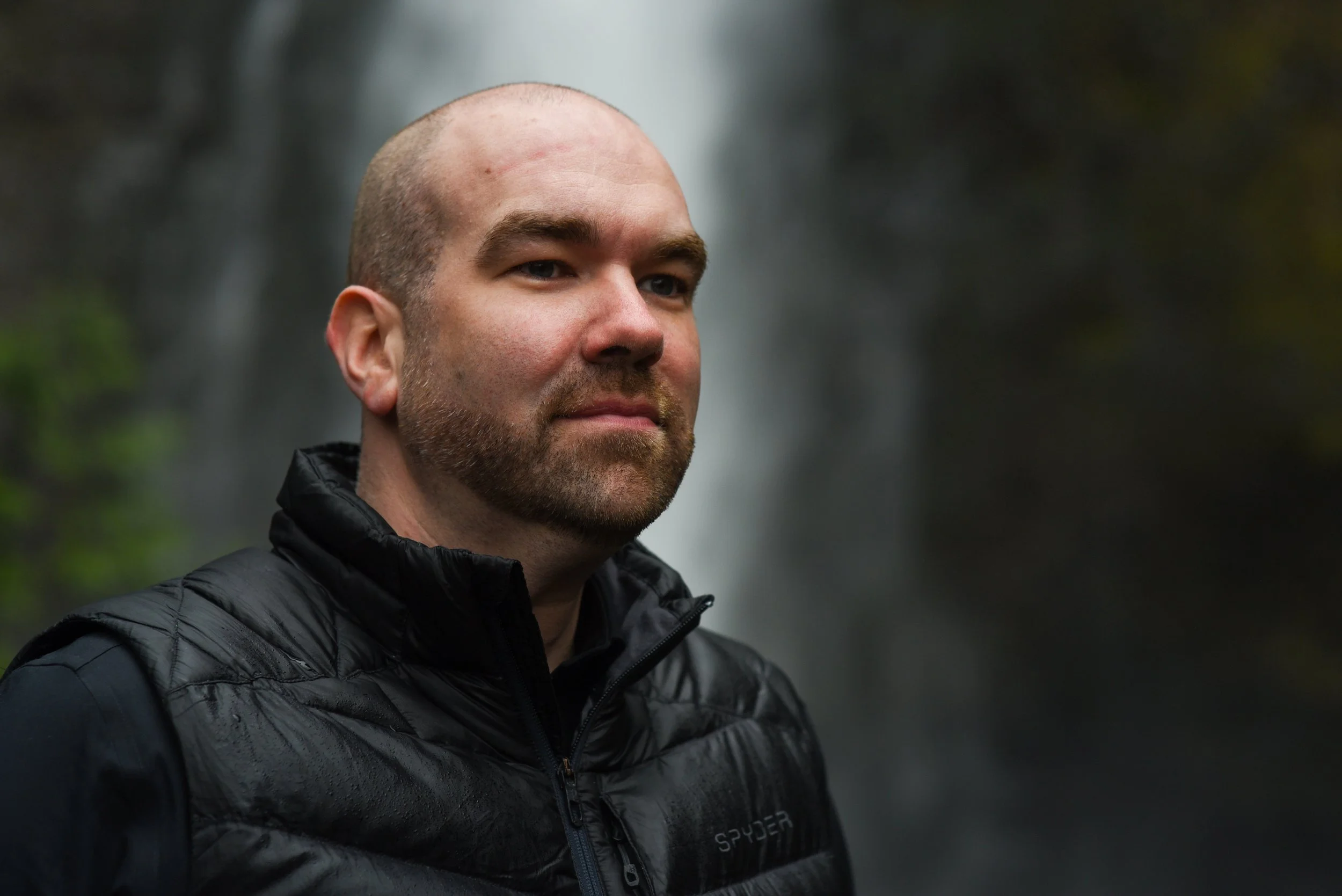 A man wearing a black jacket standing outdoors near a waterfall.