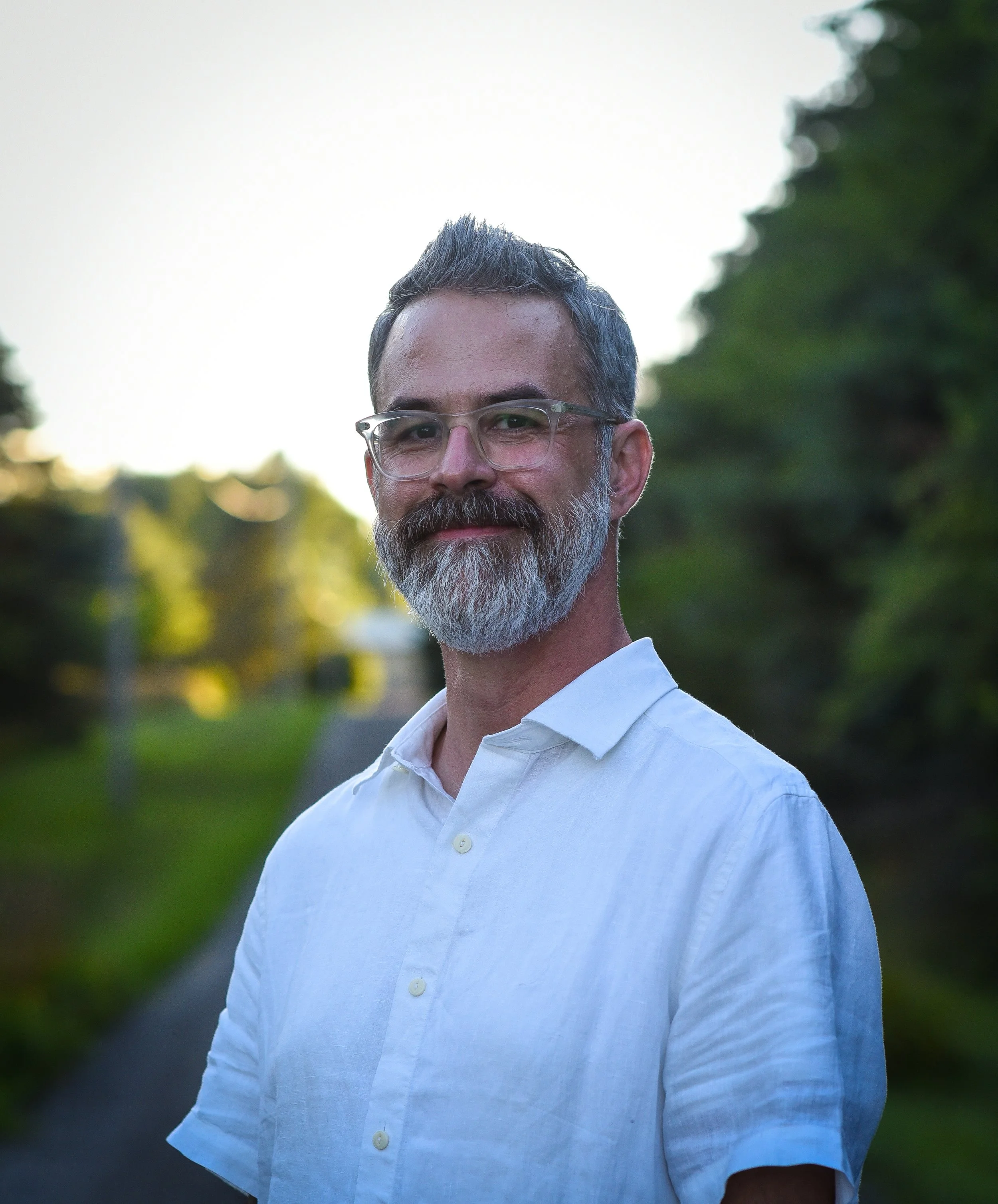 A middle-aged man with gray hair, a gray beard, and glasses, wearing a white shirt, standing outdoors with greenery and trees in the background during sunset.