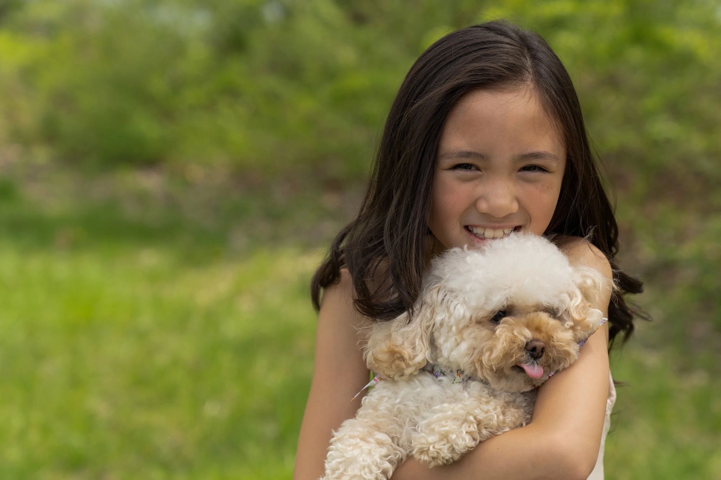 A young girl with dark hair smiling and hugging a small, curly-haired dog outdoors in a grassy area with trees in the background.