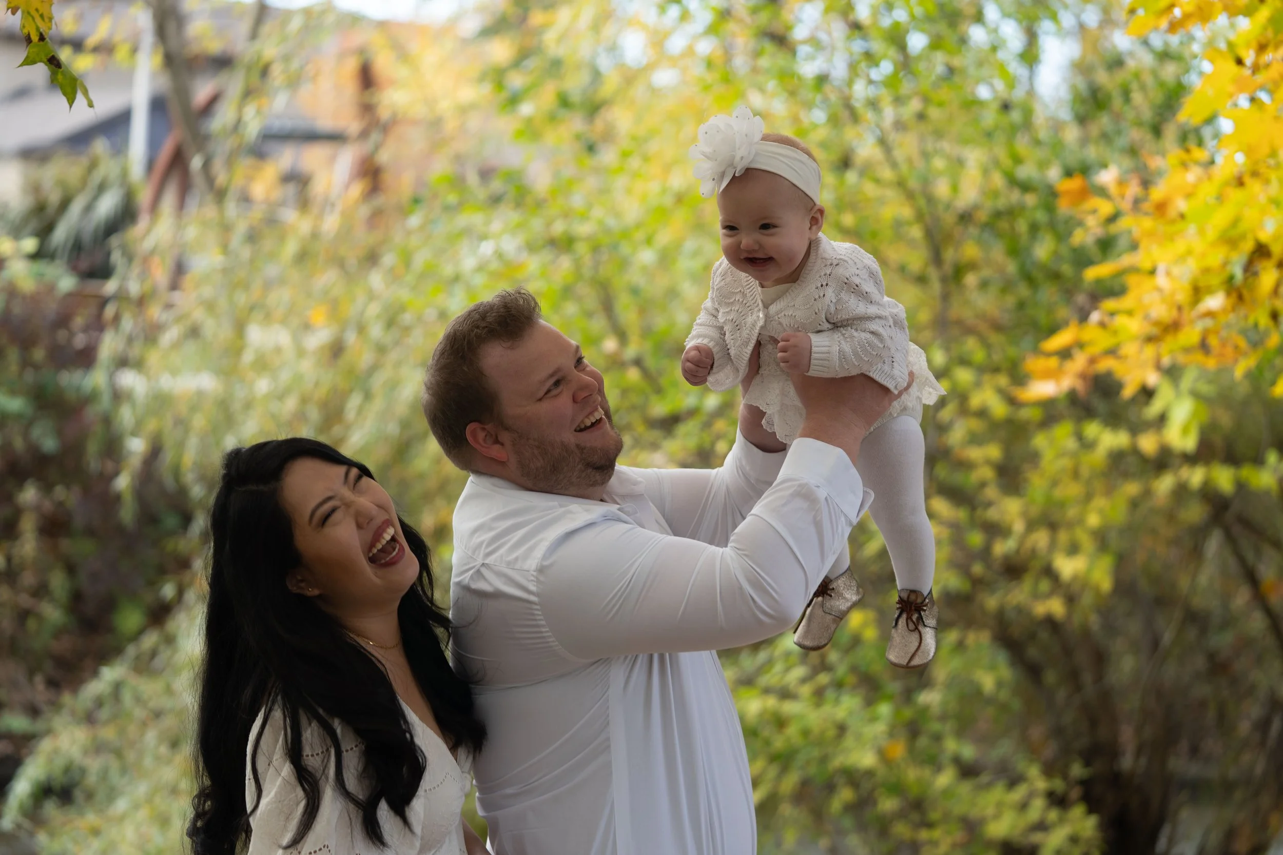 A happy family outdoors in autumn, with a man holding a smiling baby girl dressed in white, and a woman laughing nearby.