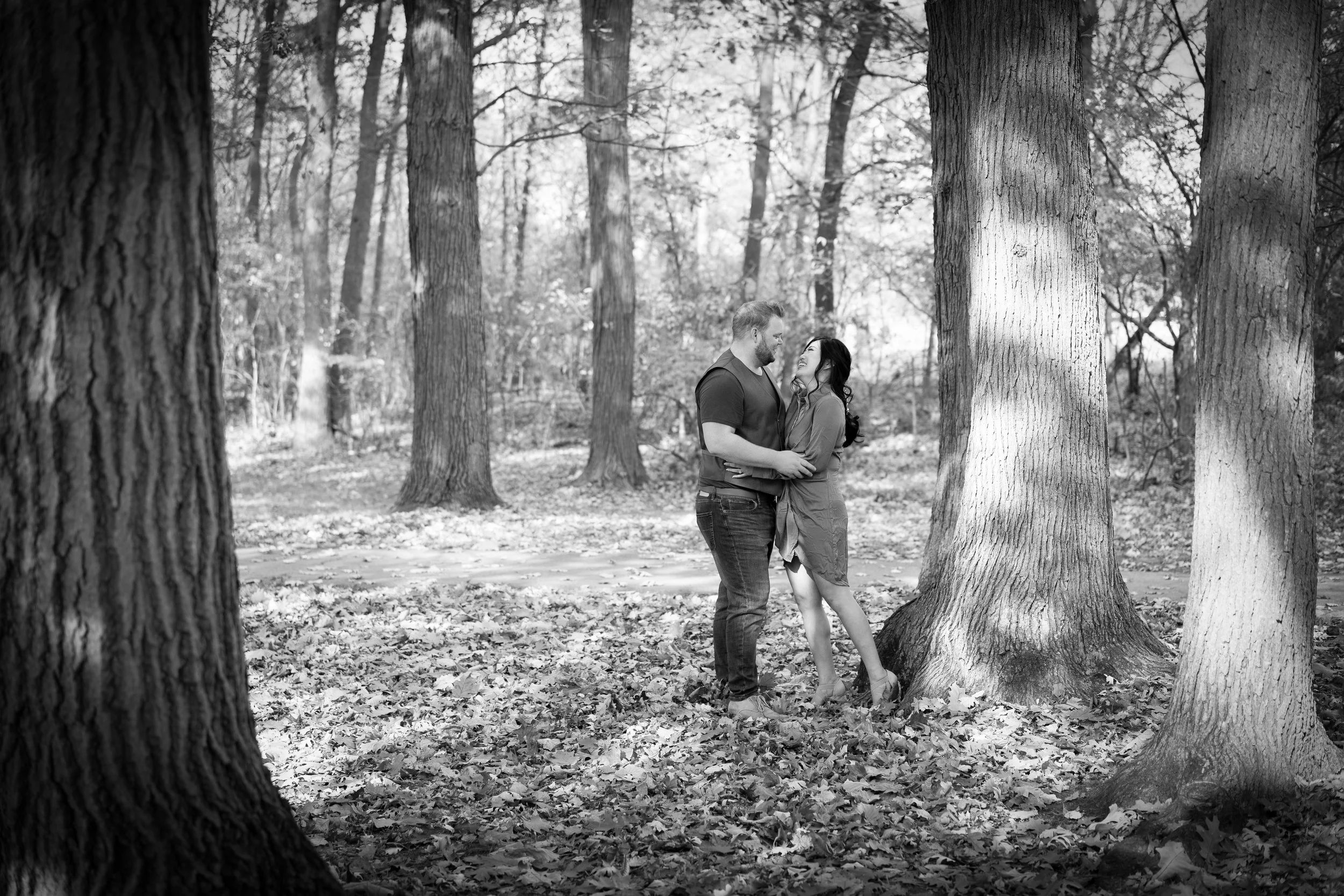 A couple standing close together in a forest, looking into each other's eyes, surrounded by tall trees and fallen leaves.