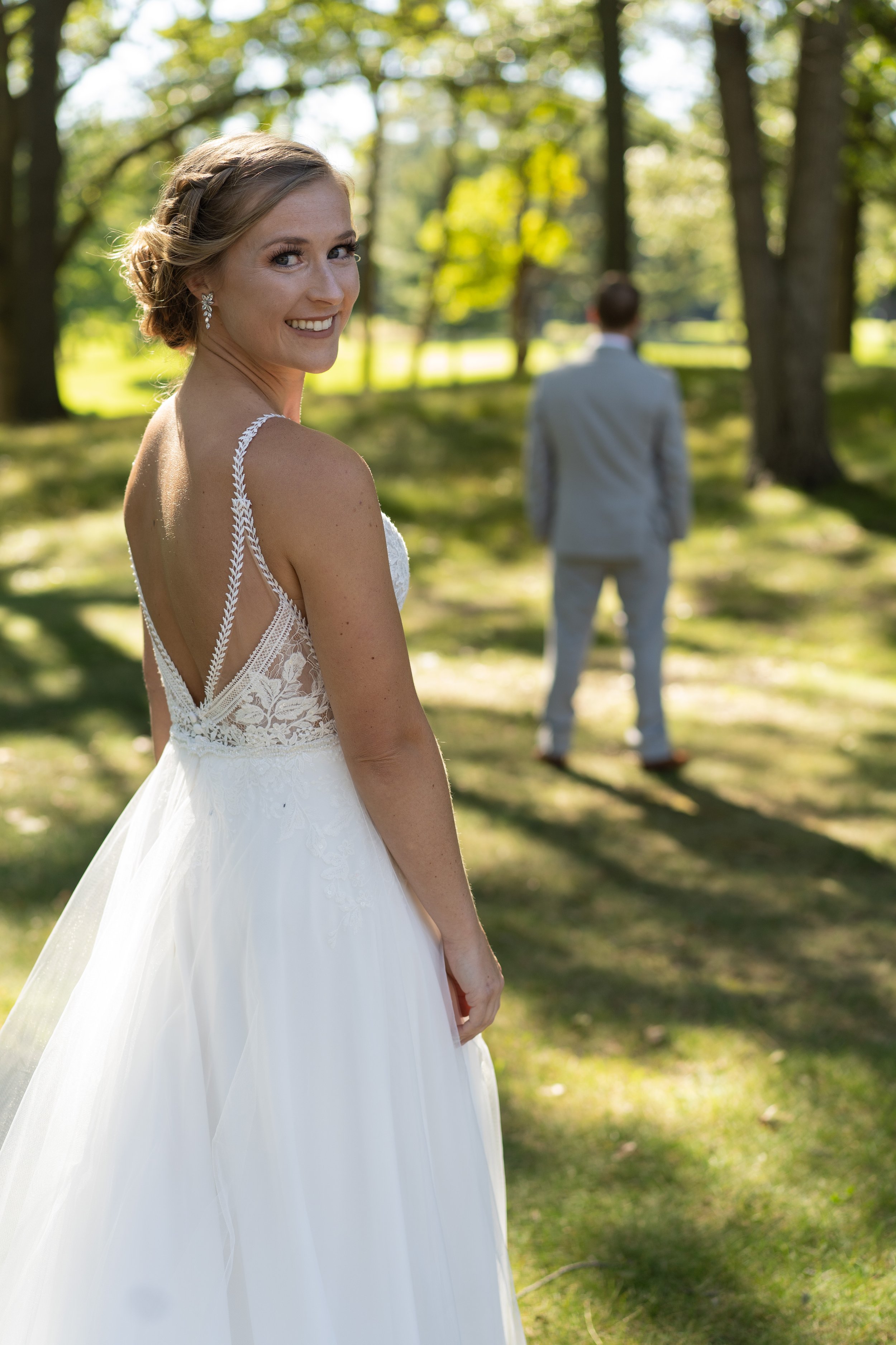 A smiling bride in a white wedding dress with lace and braid details on the straps, standing outdoors on a sunny day with trees in the background. A man in a gray suit is in the distance, walking away from her.