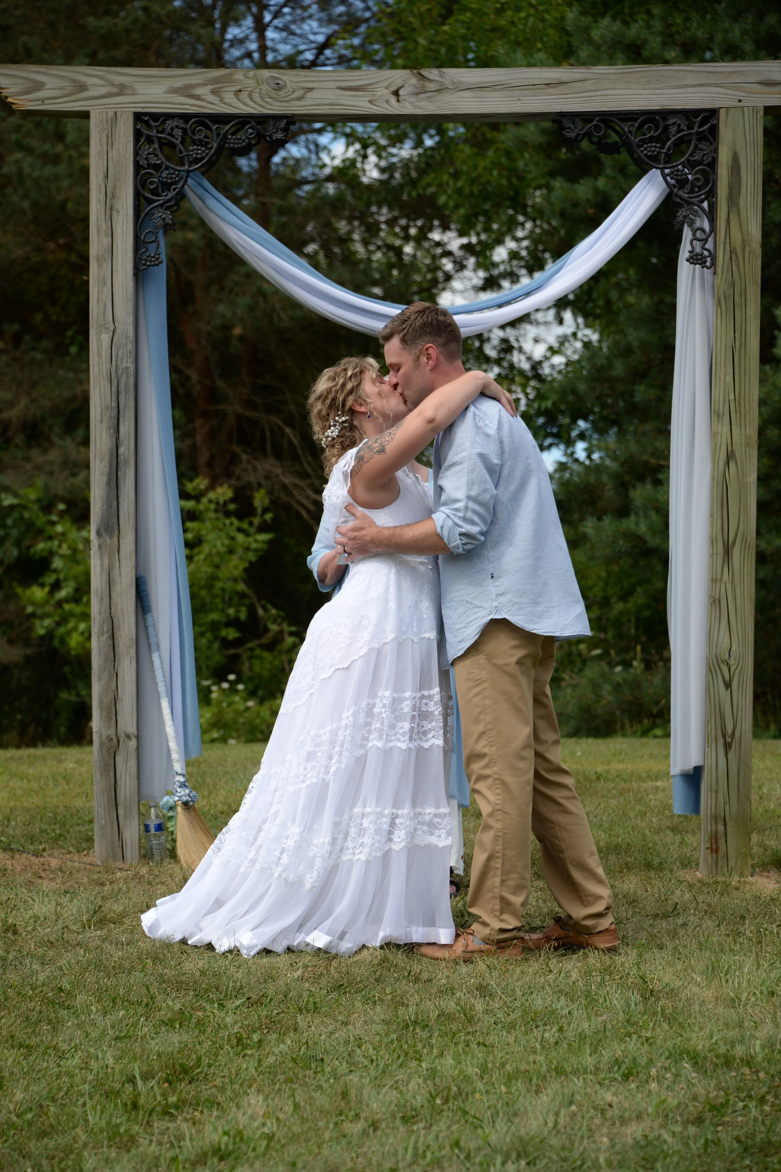 A couple kissing under an outdoor wedding arch decorated with blue and white fabric, on a grassy area with trees in the background.