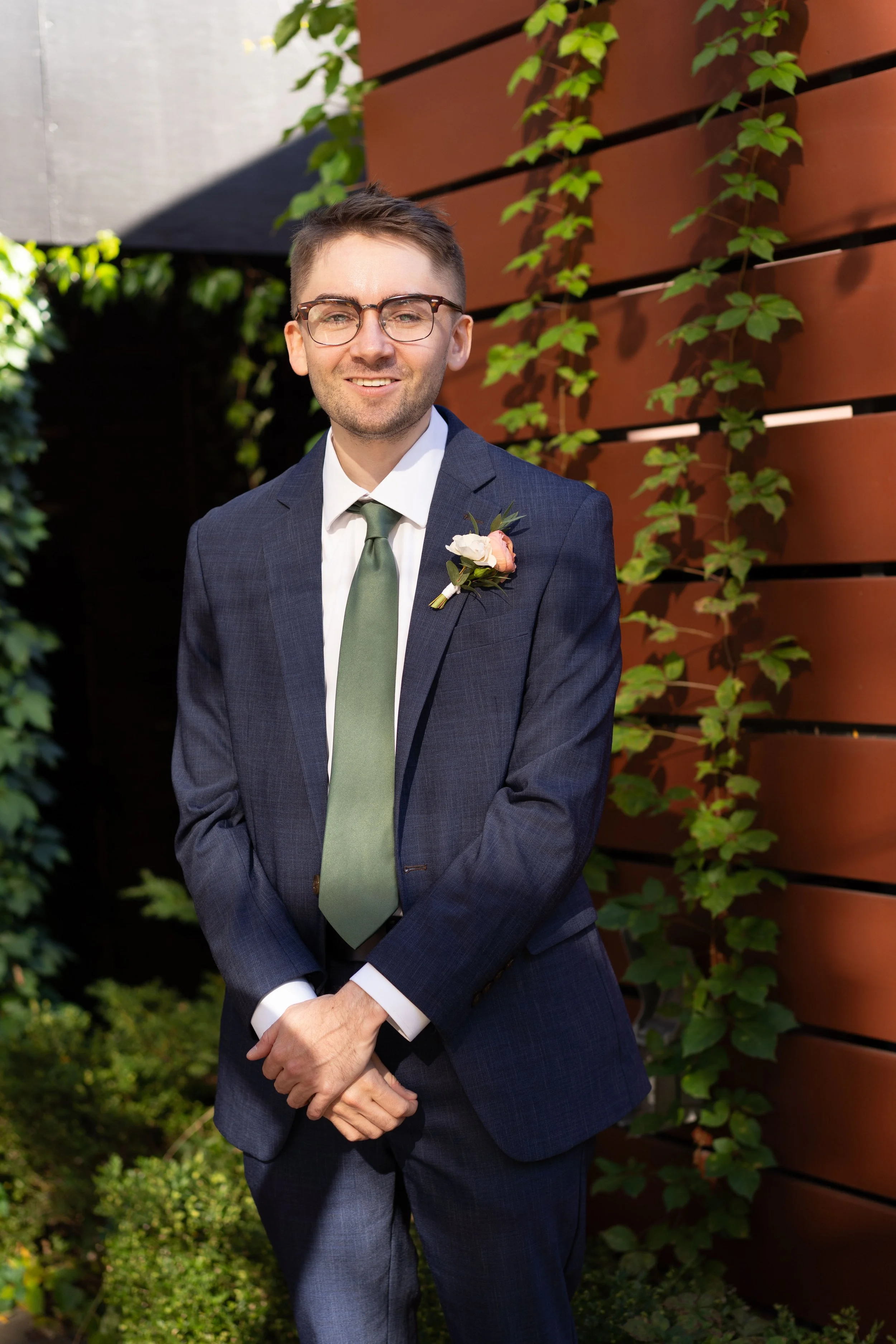 A man in a suit with glasses and a boutonniere stands outdoors against a backdrop of green plants and a wooden slat fence.