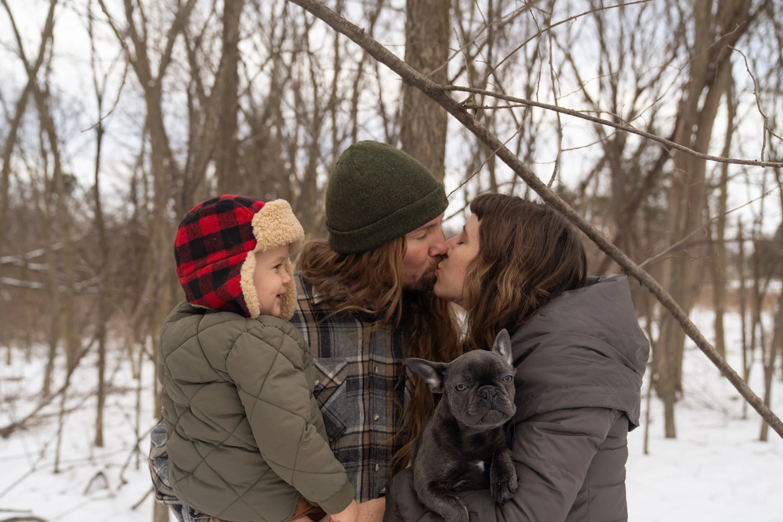 A family of three, with a dog, sharing a kiss outdoors in a snowy forest.