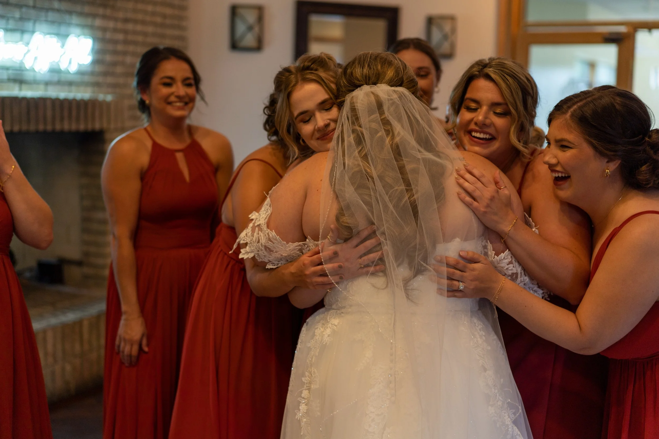 A bride in a wedding dress hugging and being hugged by her bridesmaids, all smiling and celebrating together.