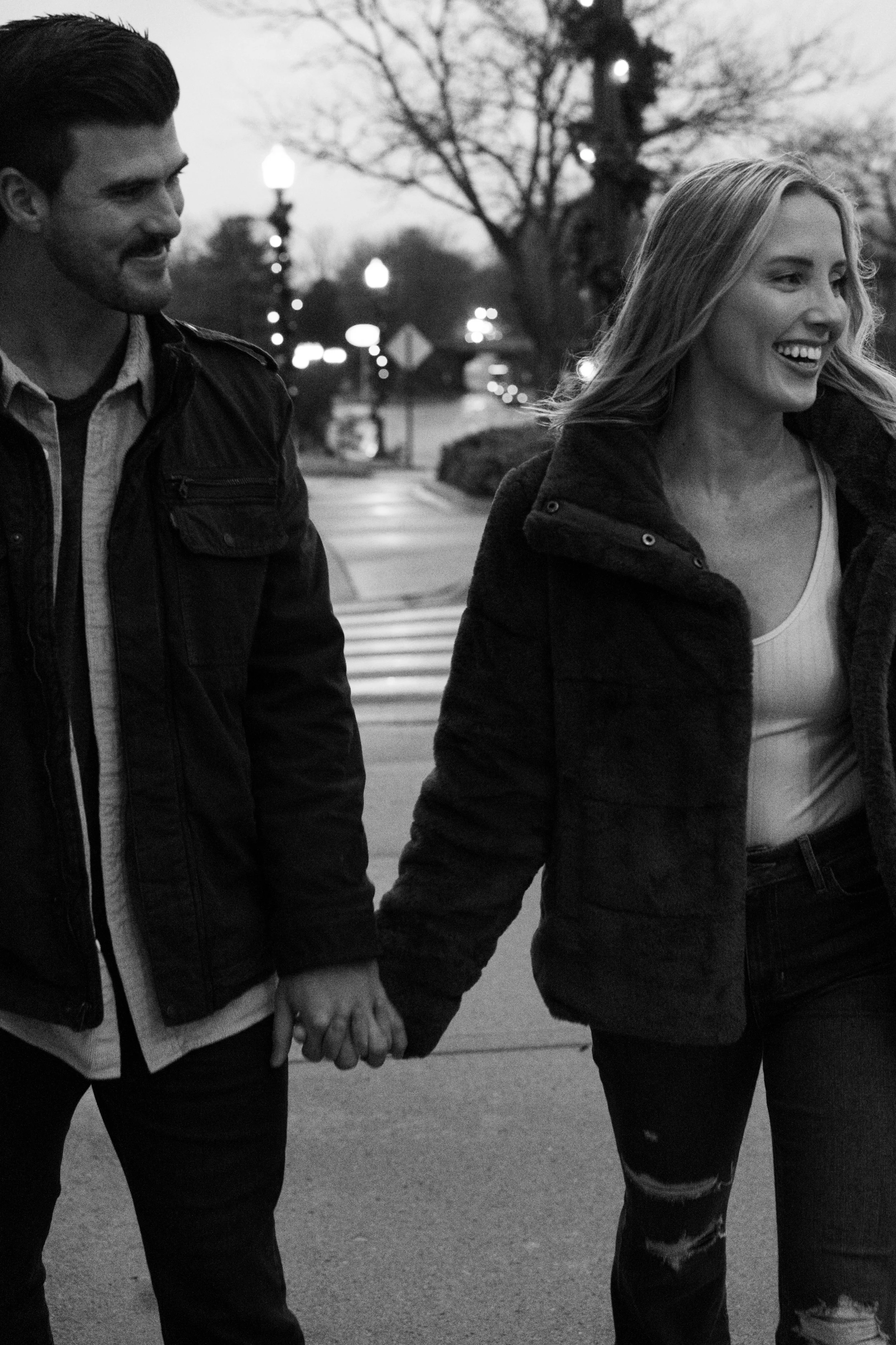 A black and white photo of a couple walking hand in hand outdoors, smiling. The man has dark hair and a beard, wearing a dark jacket, while the woman has long blonde hair, wearing a dark jacket and a white top.