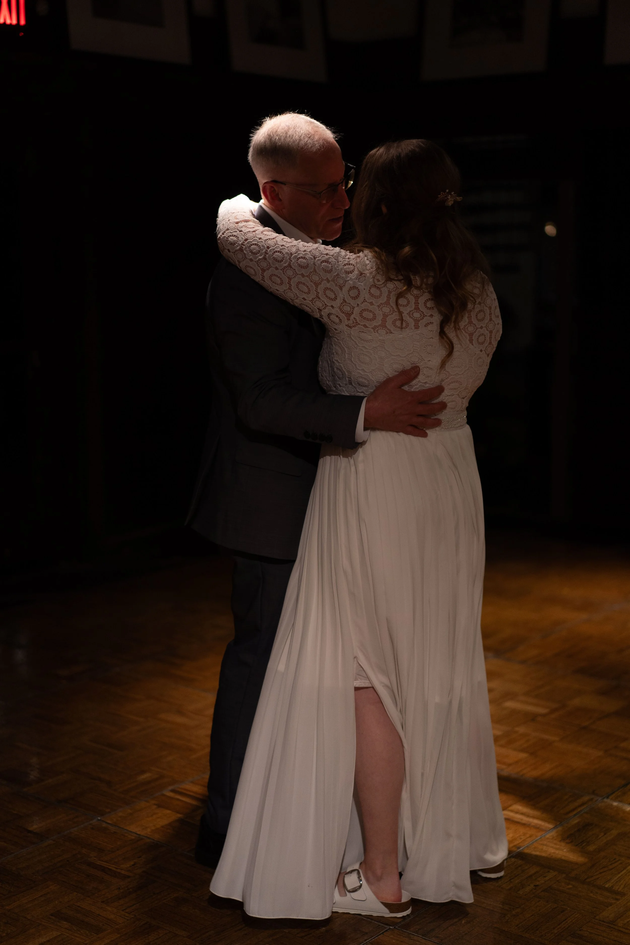 A couple dancing closely in a dimly lit room, with the woman in a white dress and the man in a dark suit.