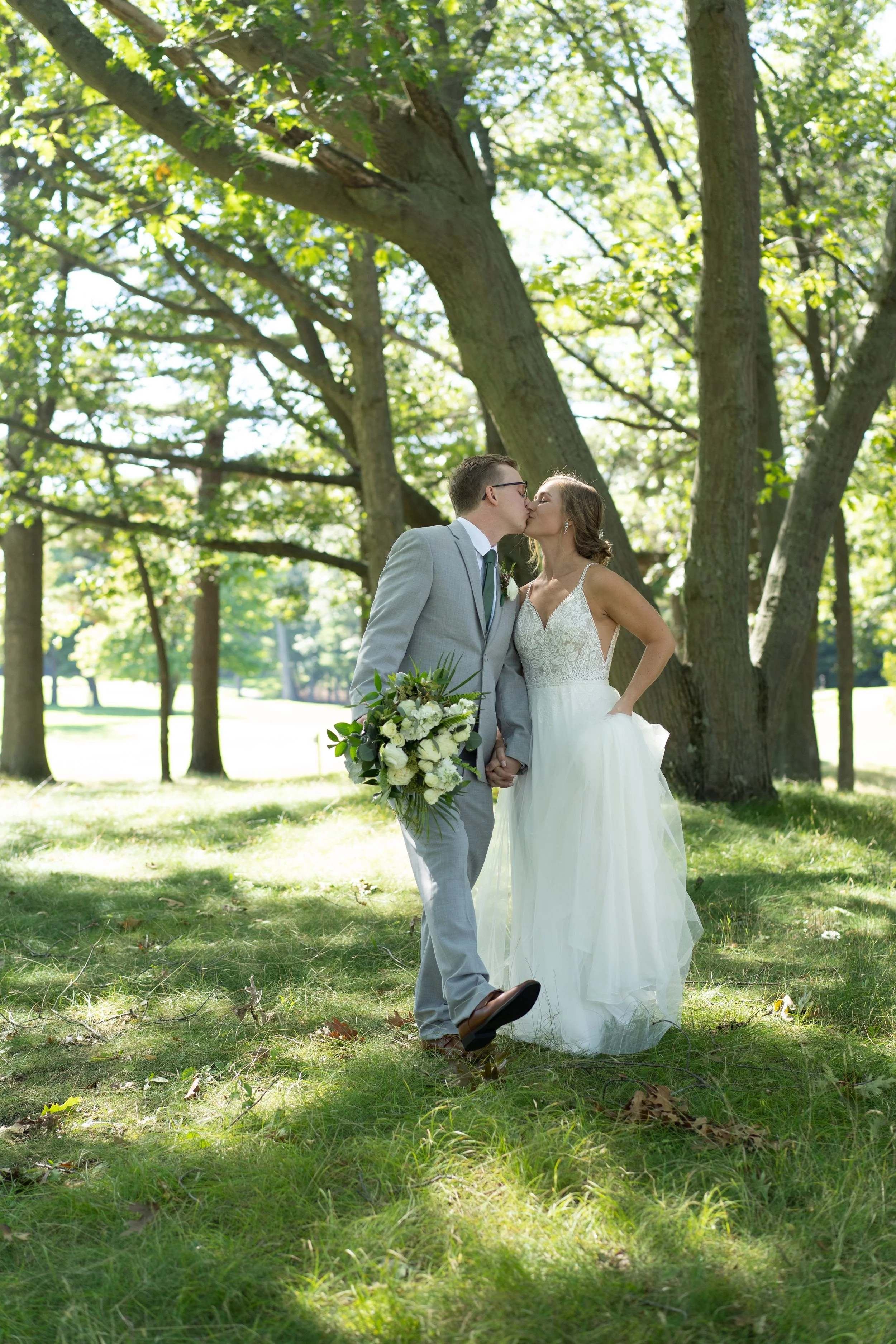 A bride and groom kiss in a park, holding hands, surrounded by tall trees and green grass on a sunny day.