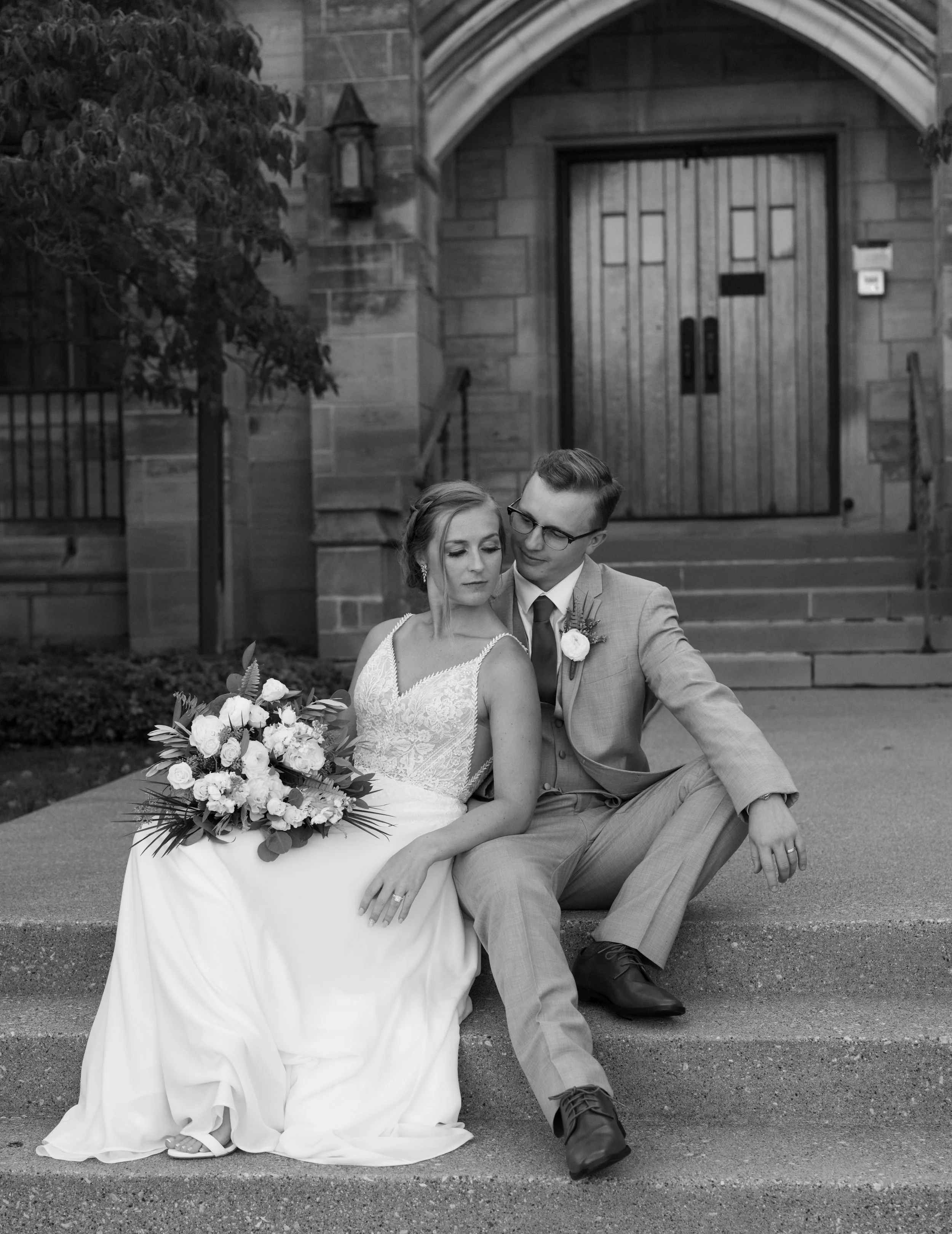 Black and white photo of a bride and groom sitting on steps outside a stone building. The bride is holding a large bouquet of flowers, and both are dressed in wedding attire.