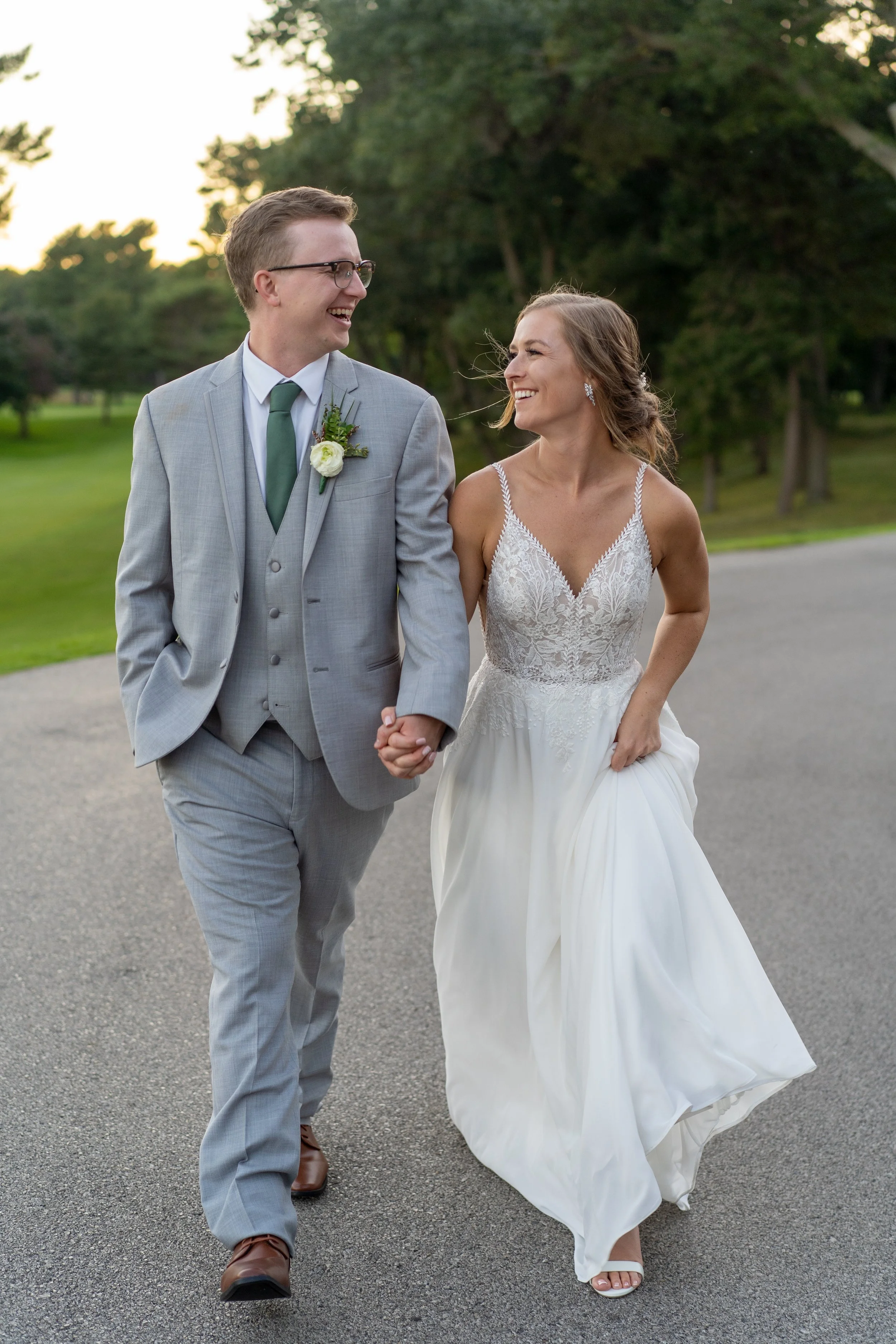 A bride and groom holding hands and walking outdoors, both smiling and looking at each other.