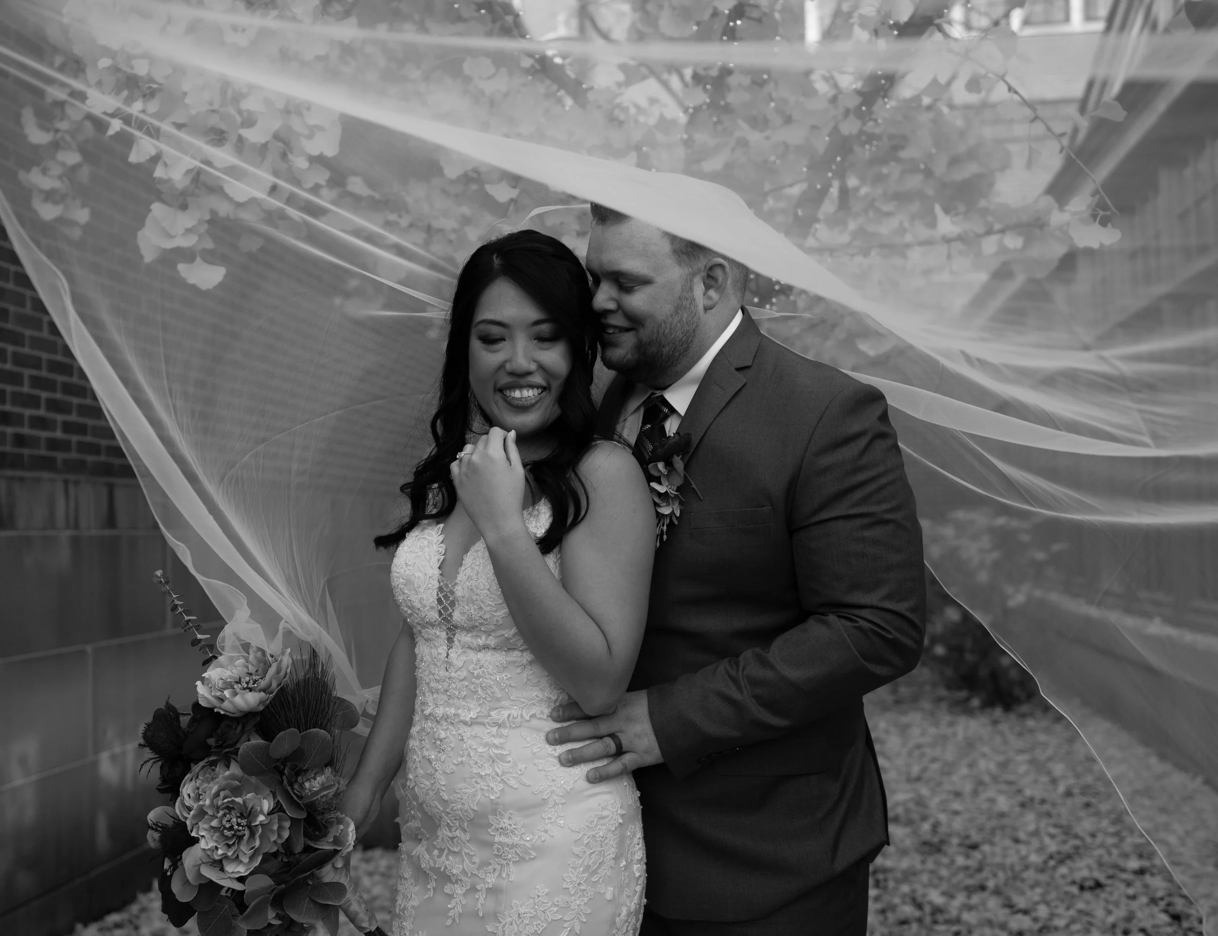 A black and white photo of a bride and groom standing close together under an umbrella. The bride is holding a bouquet of flowers, and they are smiling and looking happy.