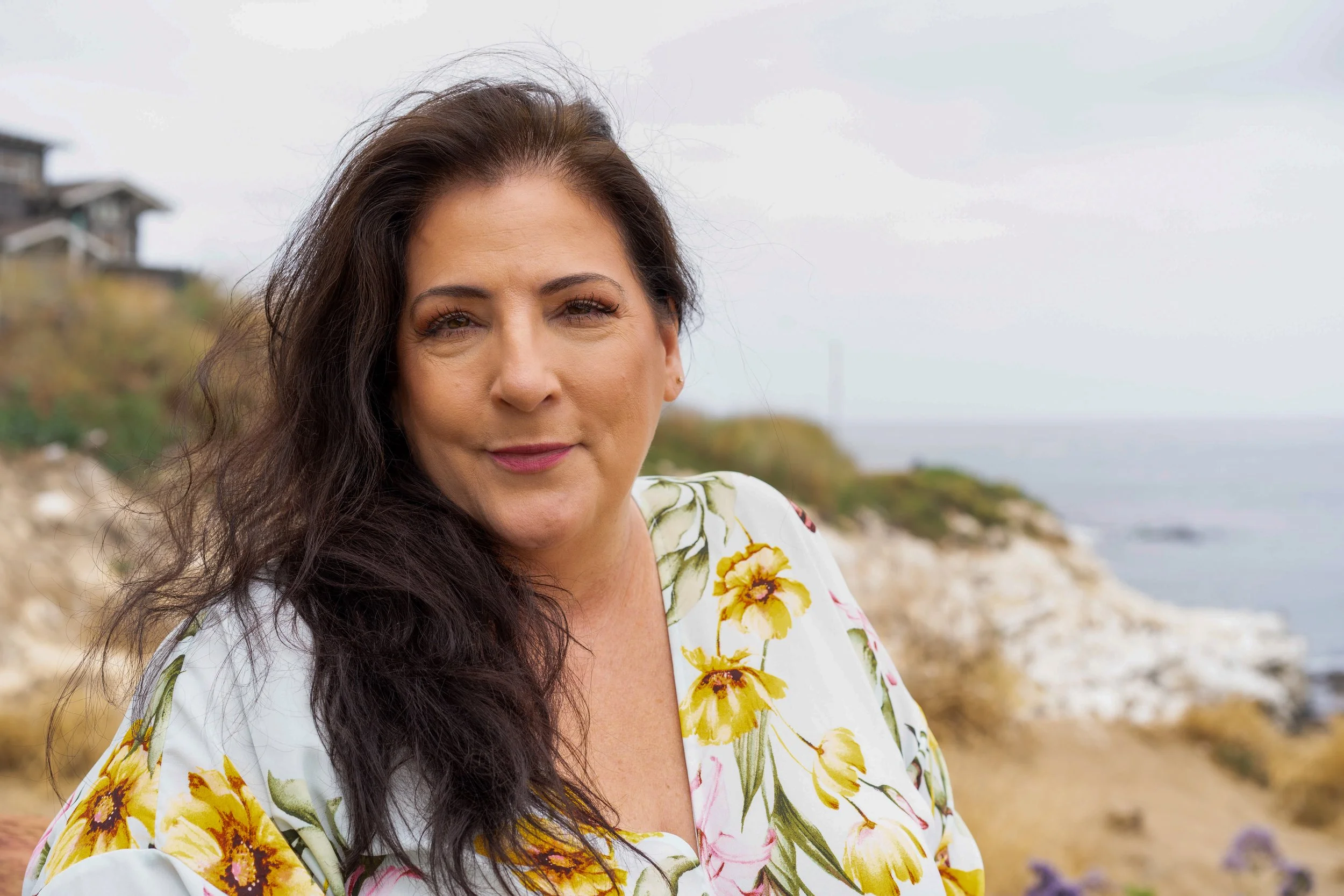 A woman with long dark hair and light makeup, wearing a white dress with a yellow floral pattern, standing outdoors near the coast with rocks, sand, and greenery in the background, under an overcast sky.