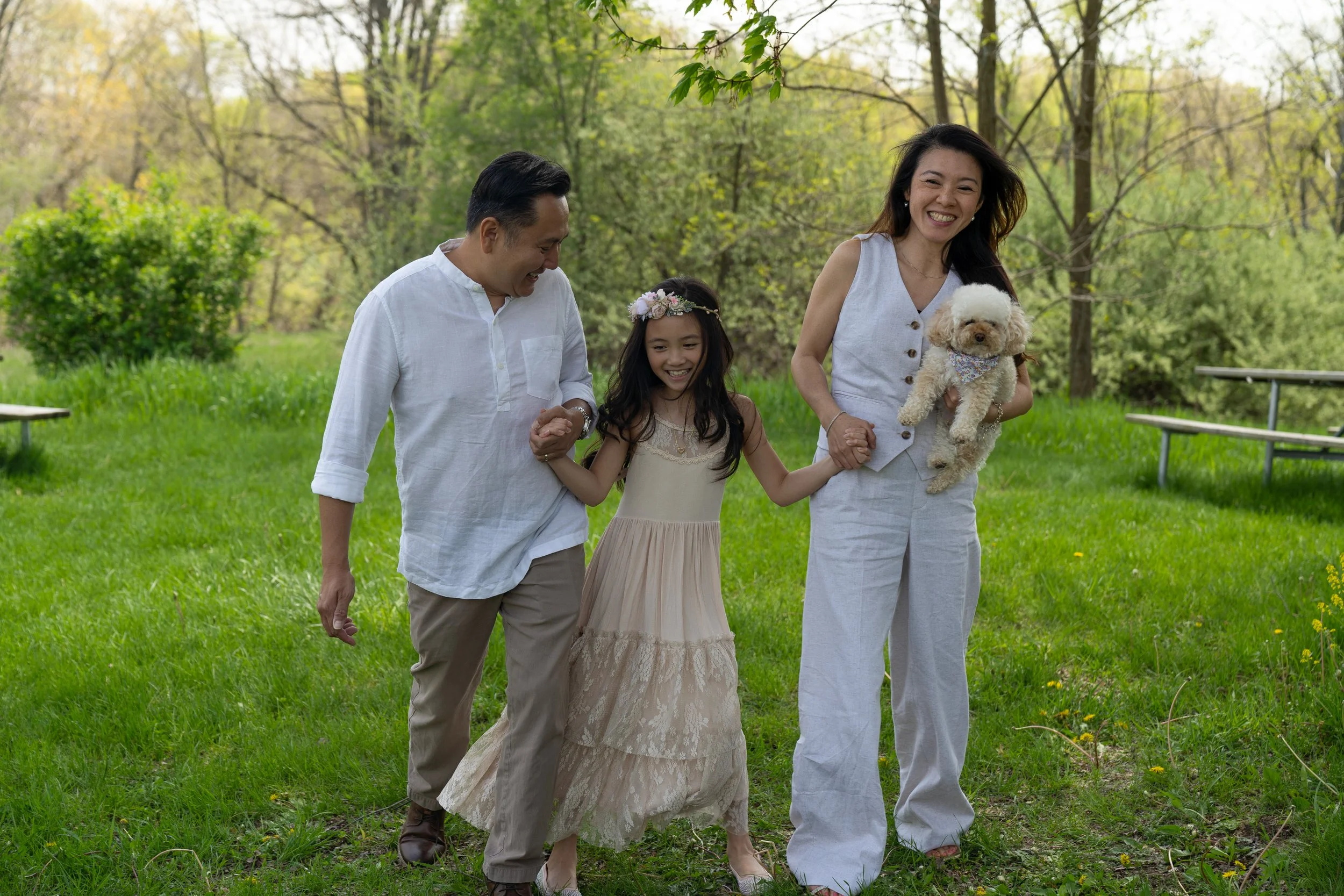 A family of four, including a man, woman, young girl, and a small dog, enjoying a walk in a park on a sunny day. They are smiling, holding hands, and dressed in light, casual clothing.