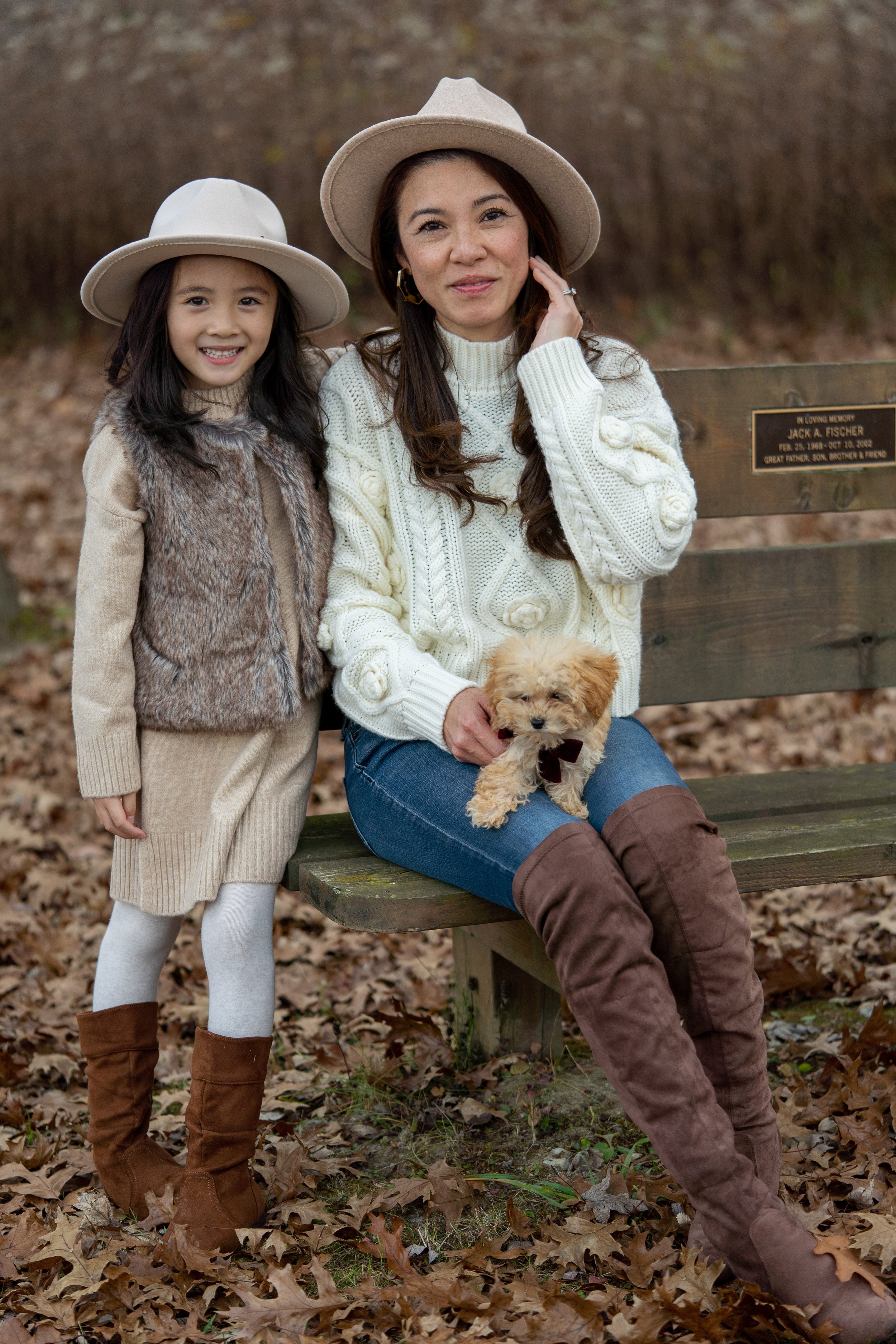 A woman and a young girl sitting on a park bench with a small puppy, all wearing fall outfits and hats, surrounded by fallen leaves.