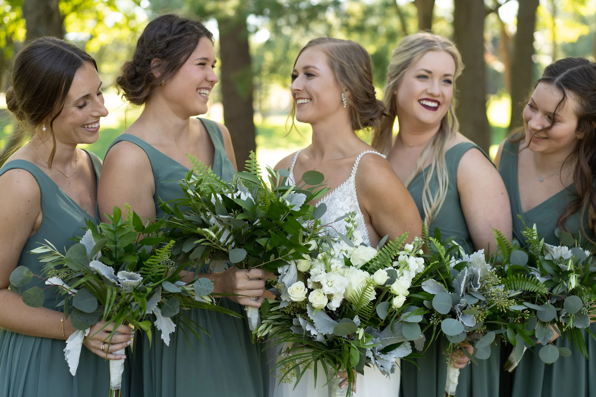 A bride in a white dress and five bridesmaids in green dresses standing outdoors, smiling and holding large bouquets of greenery and white flowers.