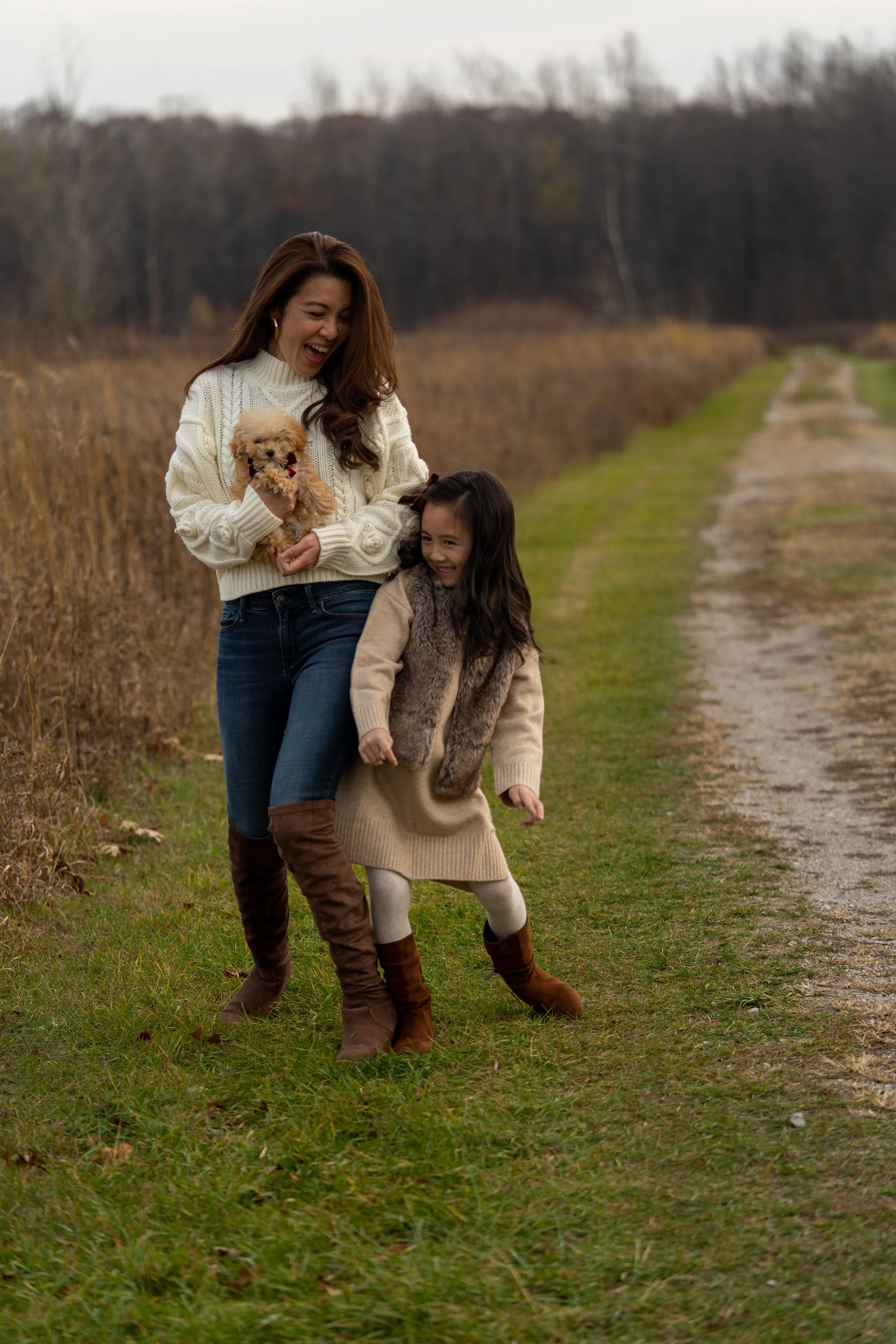 A woman and a young girl walking outdoors on a grassy path, laughing and holding a small dog, in a rural area with trees and fields in the background on a cloudy day.