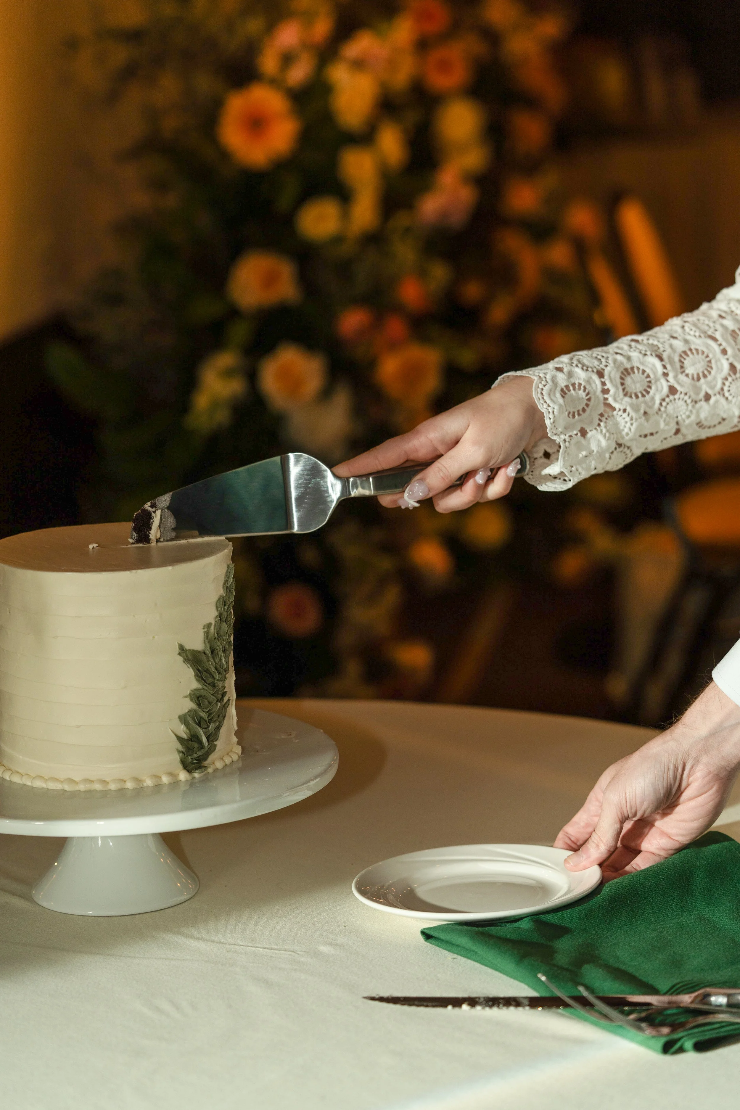 A person slicing a cake with a cake server. The cake is decorated with green leaves. The person is wearing a white lace sleeve. The background has a blurred bouquet of flowers.