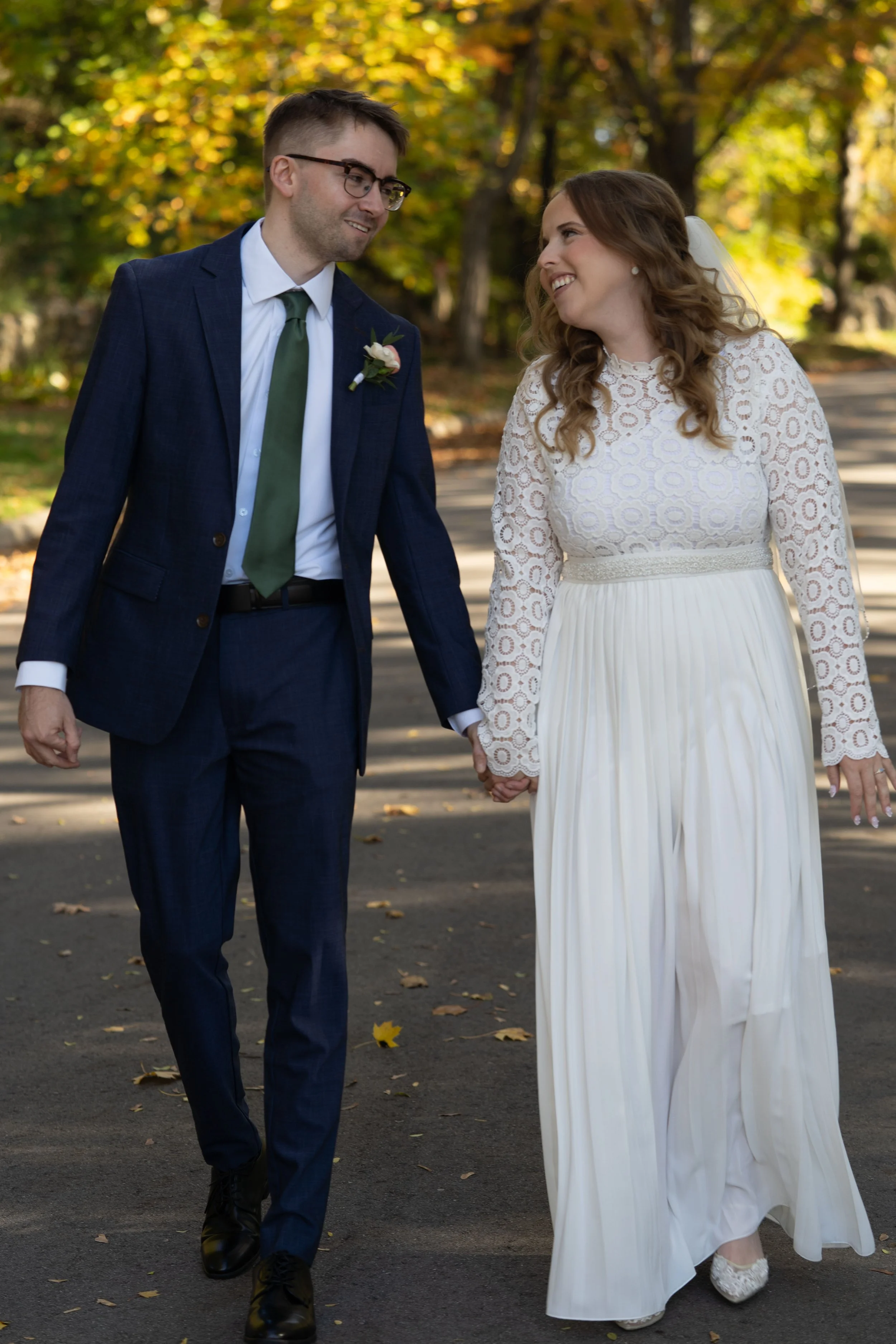 A newlywed couple holding hands and walking outdoors during fall, smiling at each other.